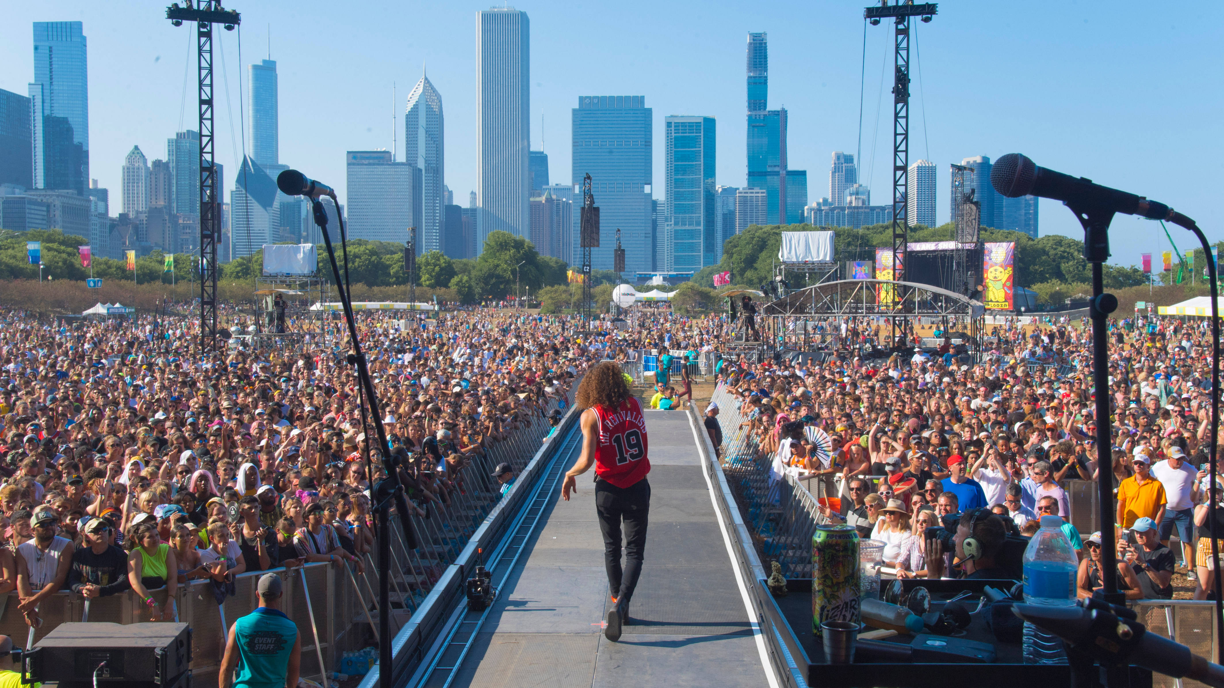 在芝加哥Lollapalooza享受夏日豔陽！ 壁紙 — 一位表演者沿著天橋走向戶外音樂會中歡呼的龐大群眾，背景是明亮的陽光下高聳的城市天際線。