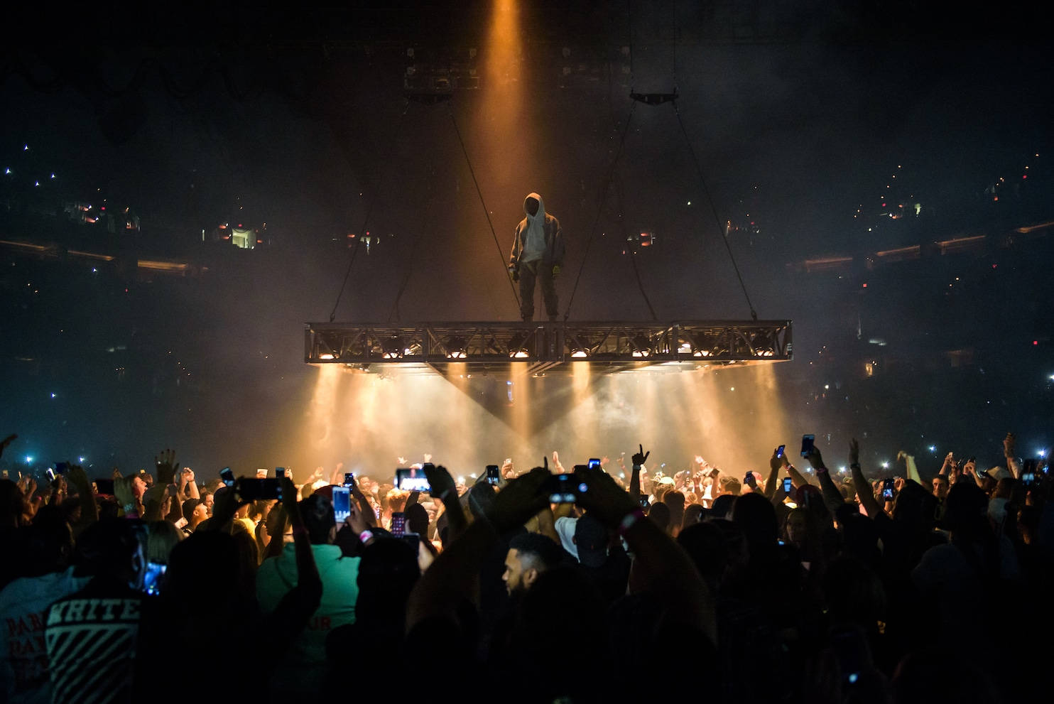 Kanye 桌布 壁紙 — 一位表演者獨自站在懸掛的舞台上，下方是咆哮的人群，演唱會沐浴在金色聚光燈下。