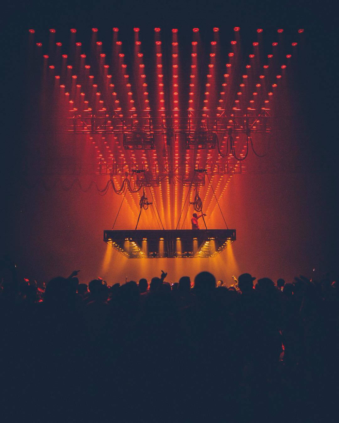 kanye west saint pablo 桌布 壁紙 — 一位表演者獨自站在懸掛的舞台上，沐浴在強烈的紅色和橙色燈光中，下方是剪影般的音樂會觀眾海洋。