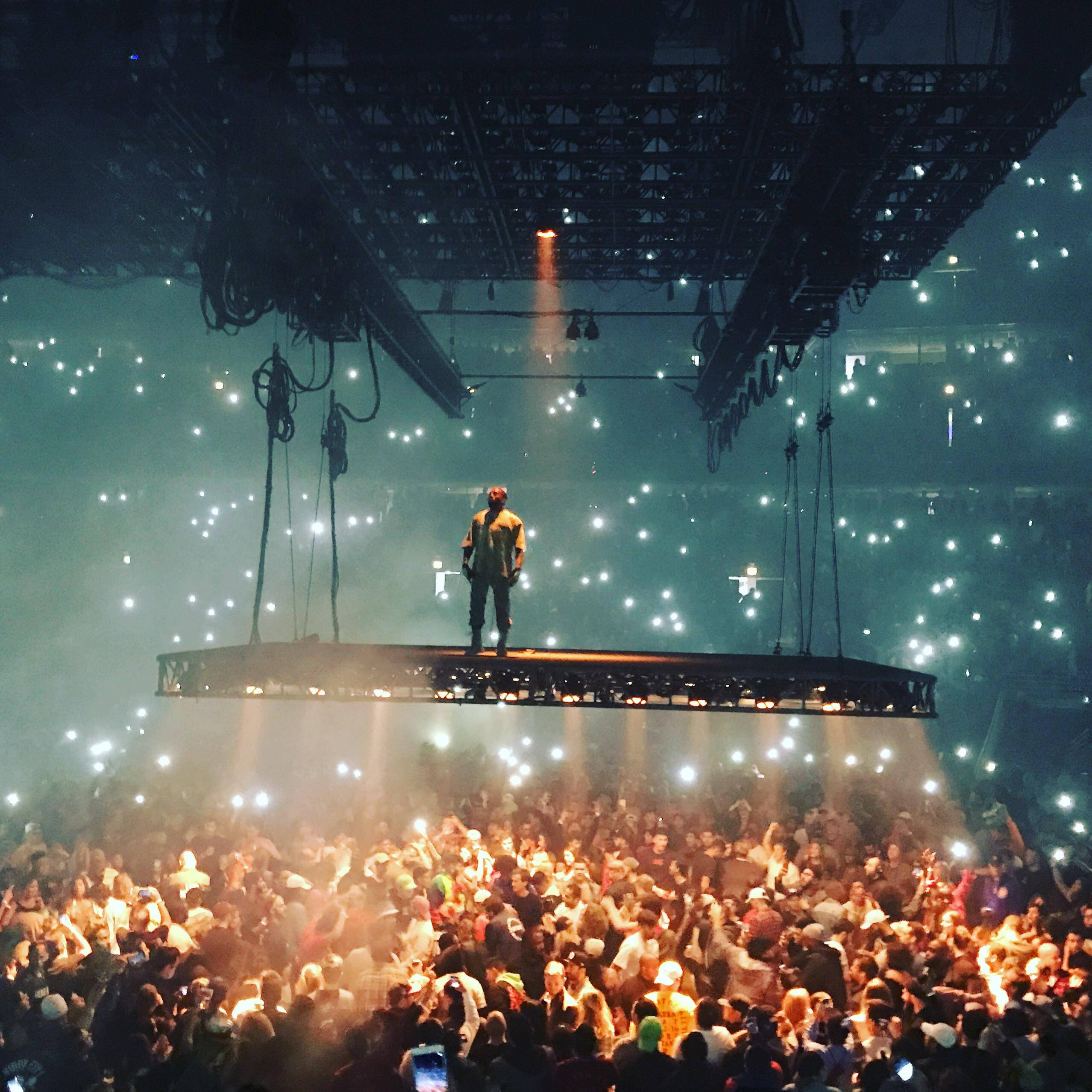 Kanye 桌布 壁紙 — 一位表演者獨自站在懸浮舞台上，下方是擠滿人的演唱會場地中一片發光手機燈海。