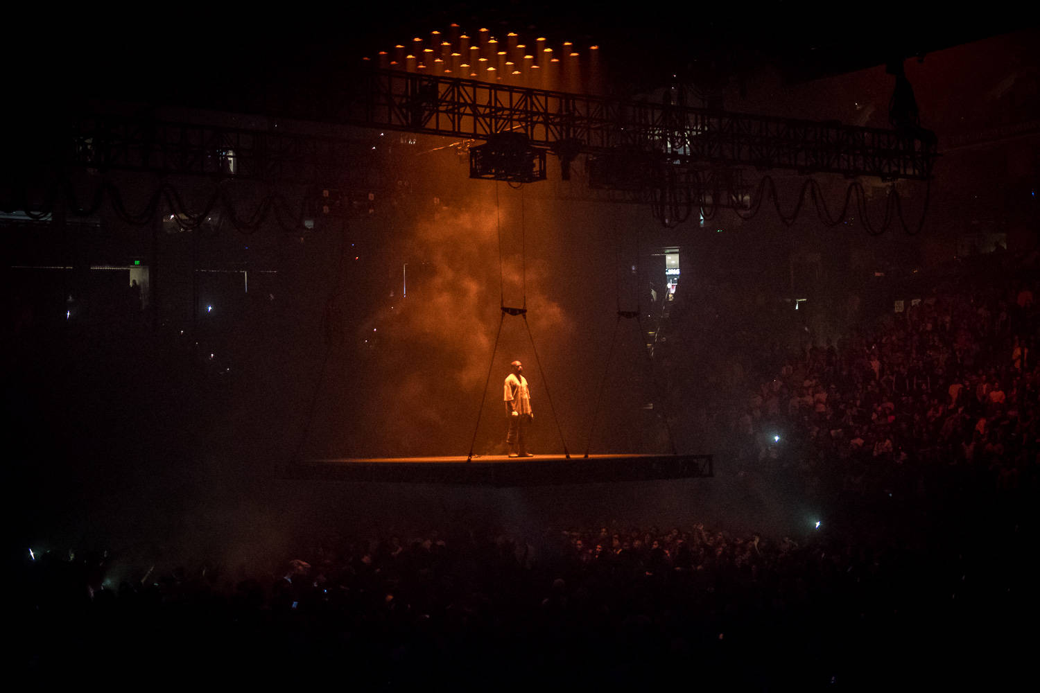 kanye west saint pablo 桌布 壁紙 — 一位表演者獨自站在懸浮舞台上，沐浴在溫暖的橙色燈光中，下方是音樂會場地中黑暗的人群。