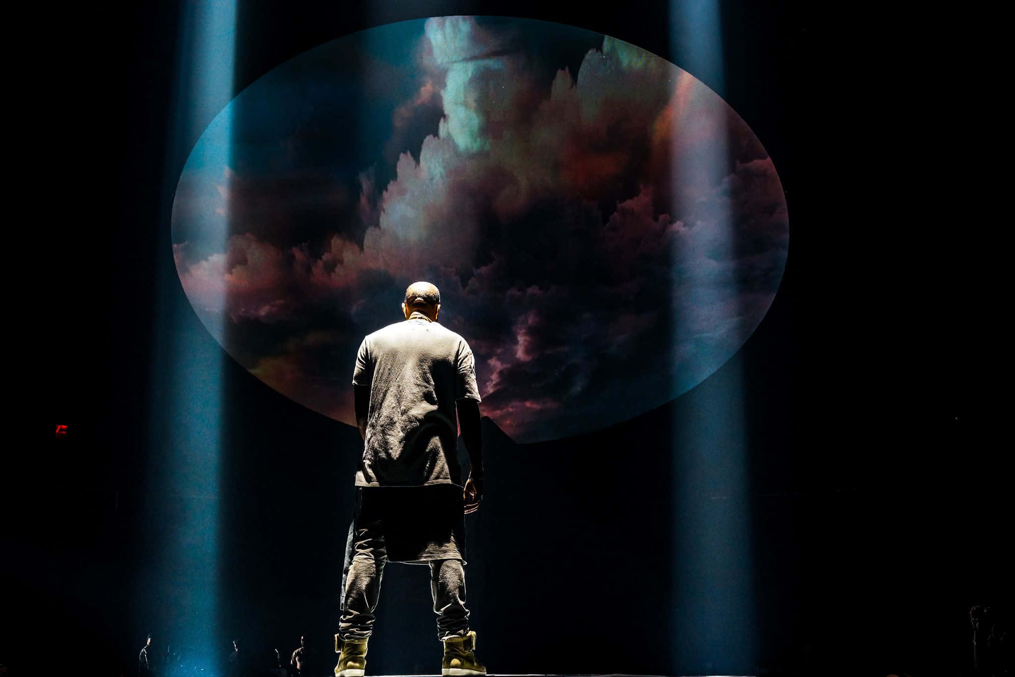 Kanye West Yeezus 桌布 壁紙 — 一位表演者獨自站在漆黑的舞台上，沐浴在藍色聚光燈下，面對一個顯示旋轉宇宙雲的巨型圓形螢幕。