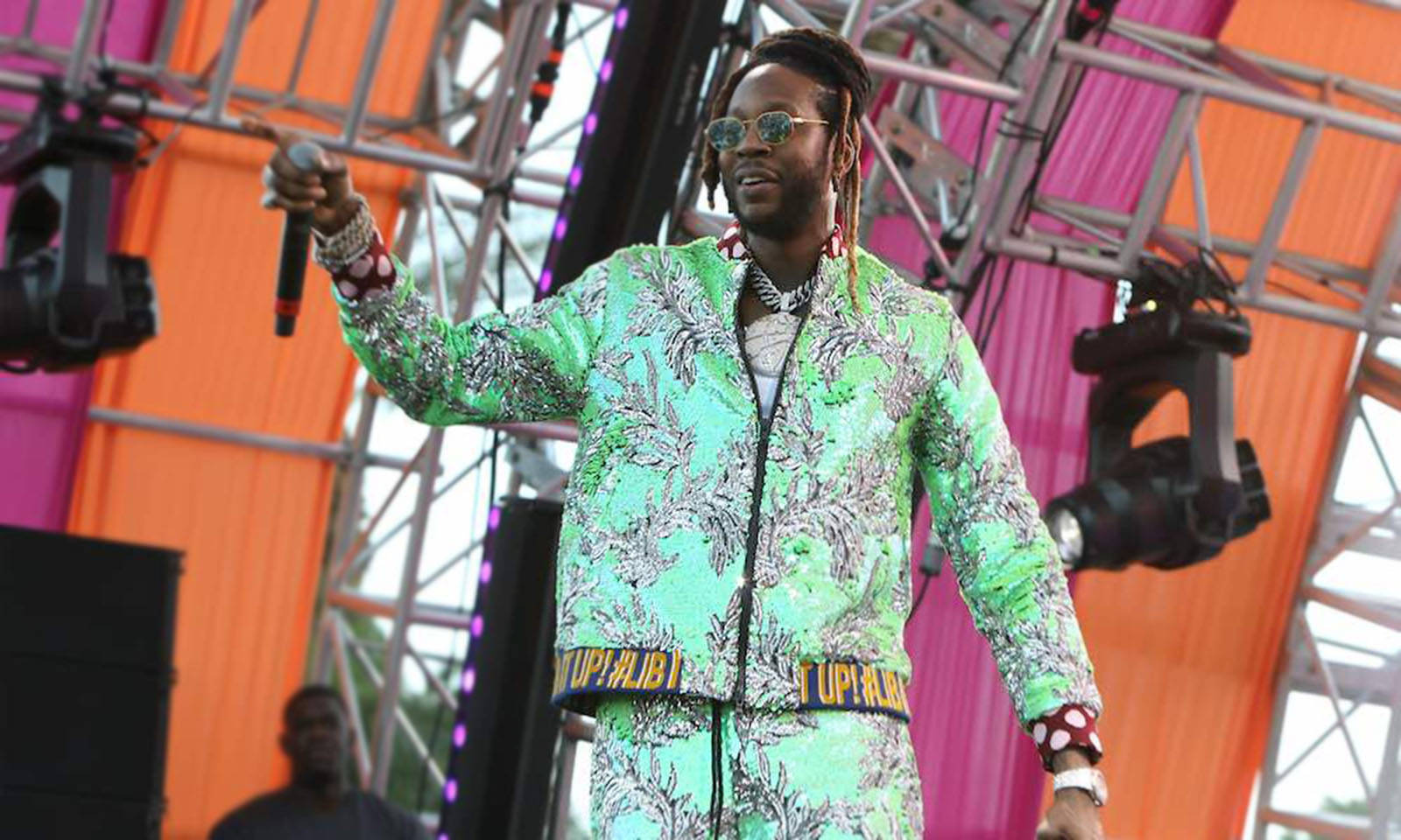 2 Chainz 桌布 壁紙 — 一位表演者穿著鮮豔的綠色圖案西裝，在色彩繽紛的戶外舞台上，對著麥克風唱歌，舞台上有金屬桁架和聚光燈。