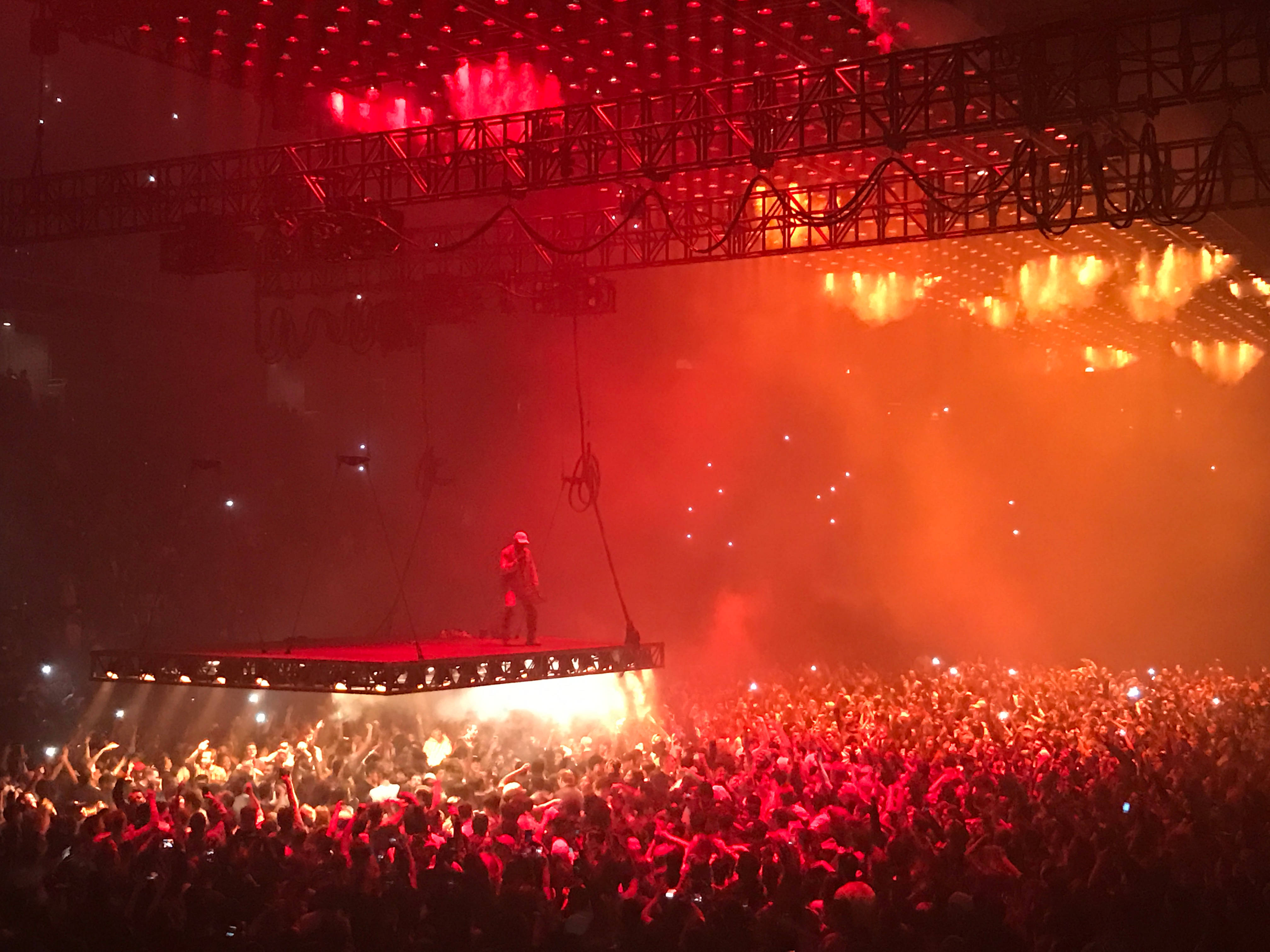 kanye west saint pablo 桌布 壁紙 — 一位表演者站在懸掛的平台上，下方是沐浴在強烈紅色舞台燈光和煙霧中的咆哮人群。