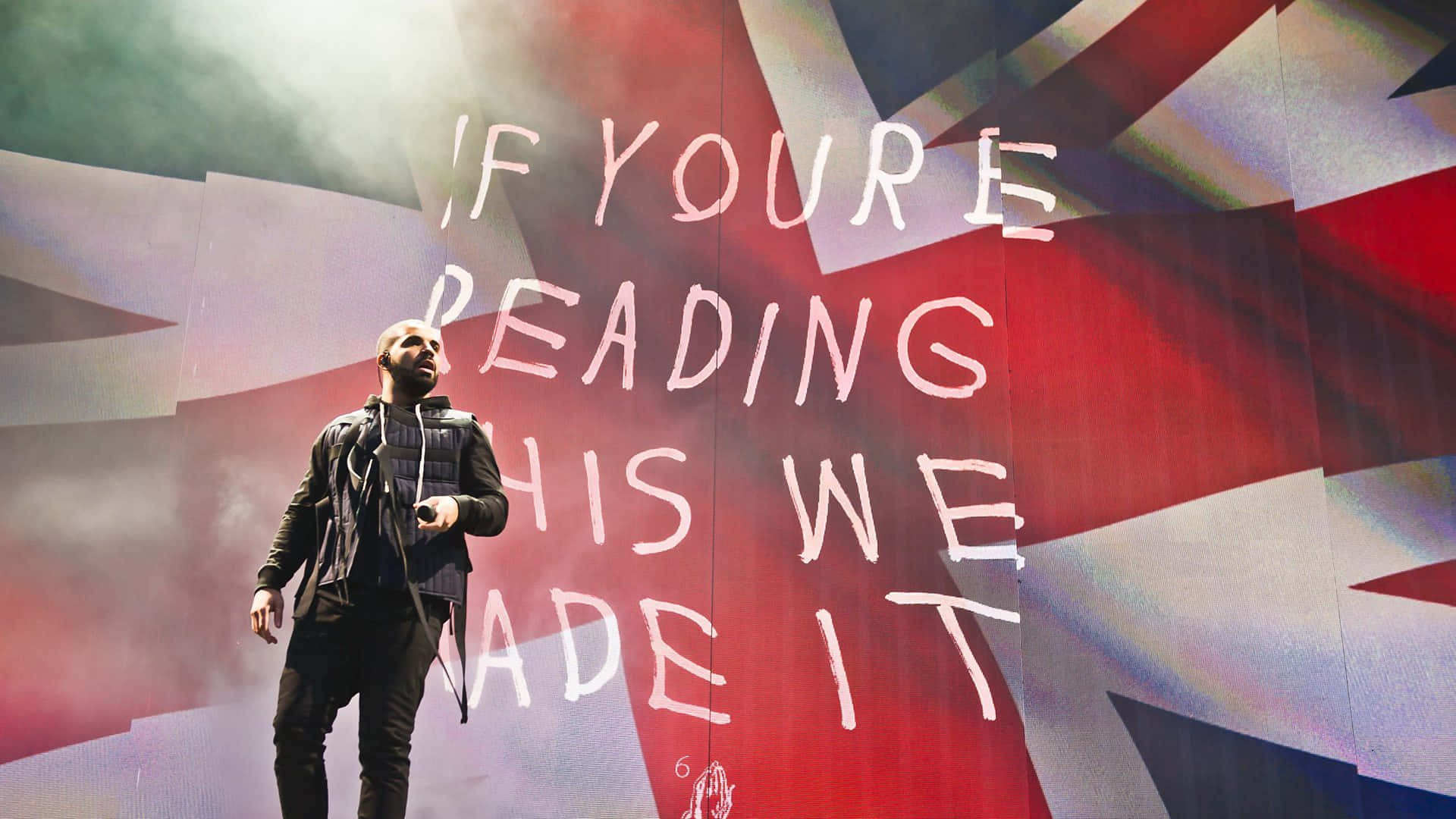 桌布 壁紙 — 一位表演者站在舞台上，背景是巨大的英國國旗，上面有「if youre reading this we made it」的白色發光字句。