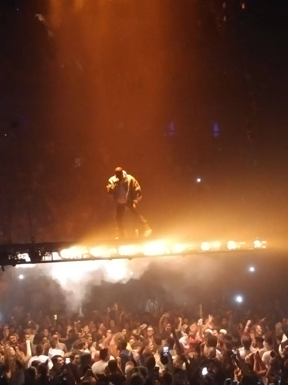 kanye west android 桌布 壁紙 — 一位表演者站在被橙色燈光籠罩的舞台上，下方是音樂會中喧鬧的人群。