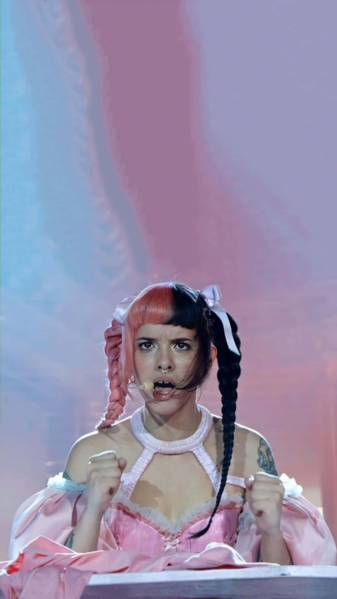Melanie Martinez 桌布 壁紙 — 一位表演者紮著粉紅和黑色辮子，身穿粉紅連衣裙，在彩色燈光下激情演唱。