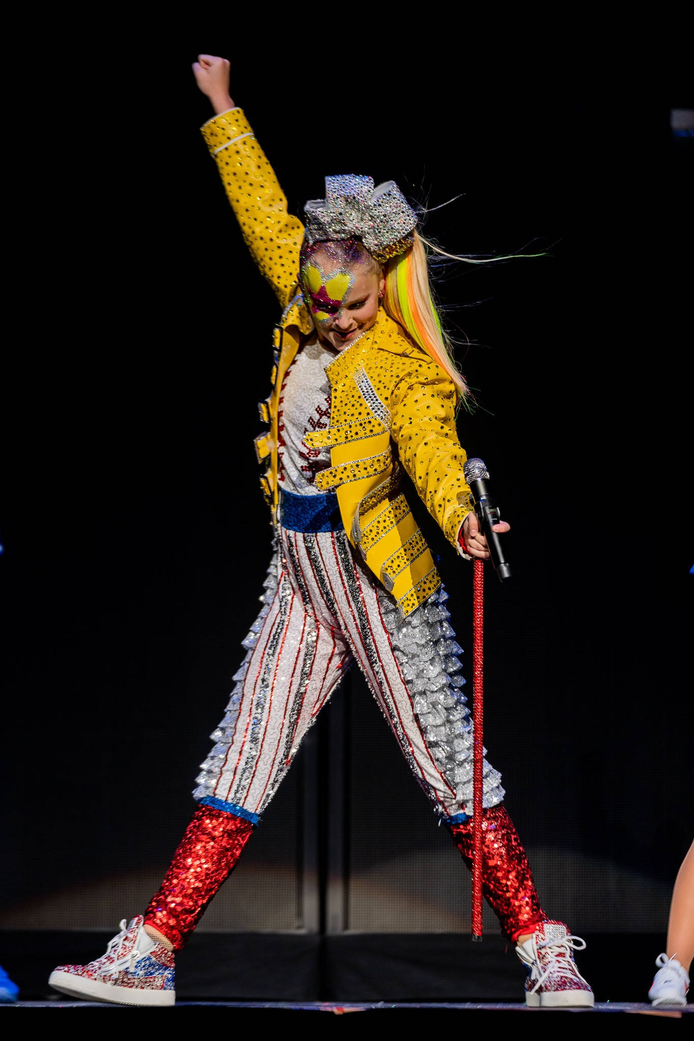 JoJo Siwa 桌布 壁紙 — 一位表演者身穿閃亮黃色外套和條紋褲，在舞台上擺出動感姿勢，一隻手臂高舉，手中拿著麥克風。