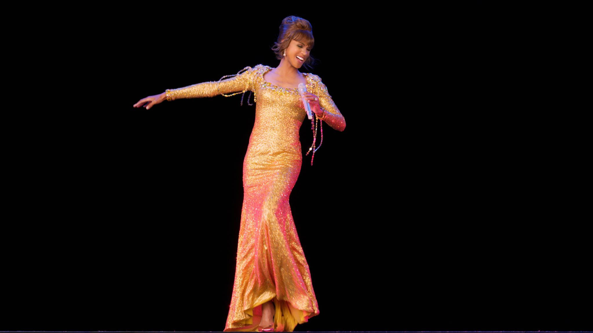 Whitney 桌布 壁紙 — 一位表演者身穿閃爍金色和粉紅色亮片長裙，微笑並伸展雙臂，背景為黑色。