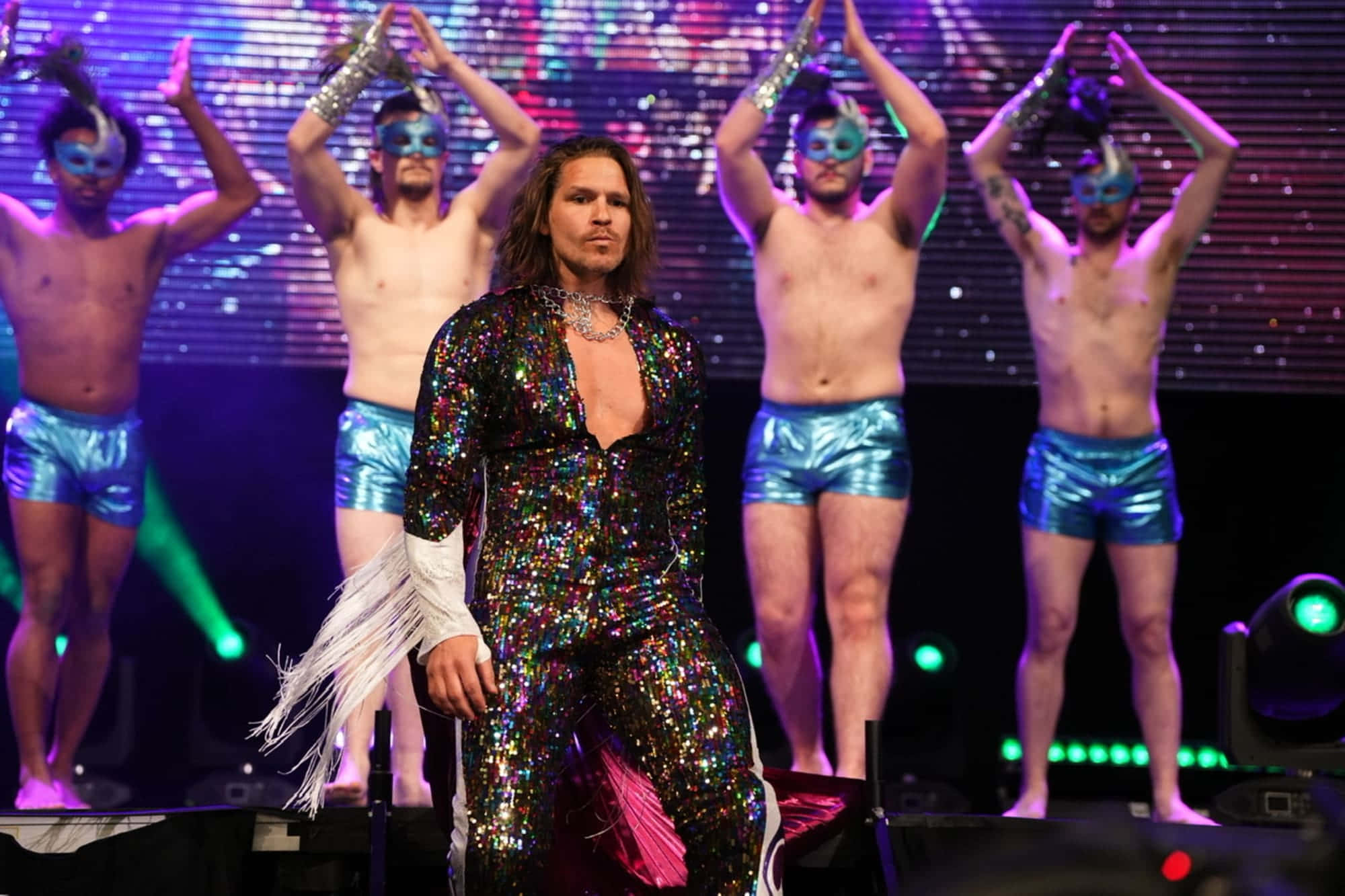 Aew 桌布 壁紙 — 一位表演者身穿閃閃發光的亮片連身衣站在舞台中央，戴著面具的伴舞在充滿活力的舞台燈光下擺出同步的姿勢。