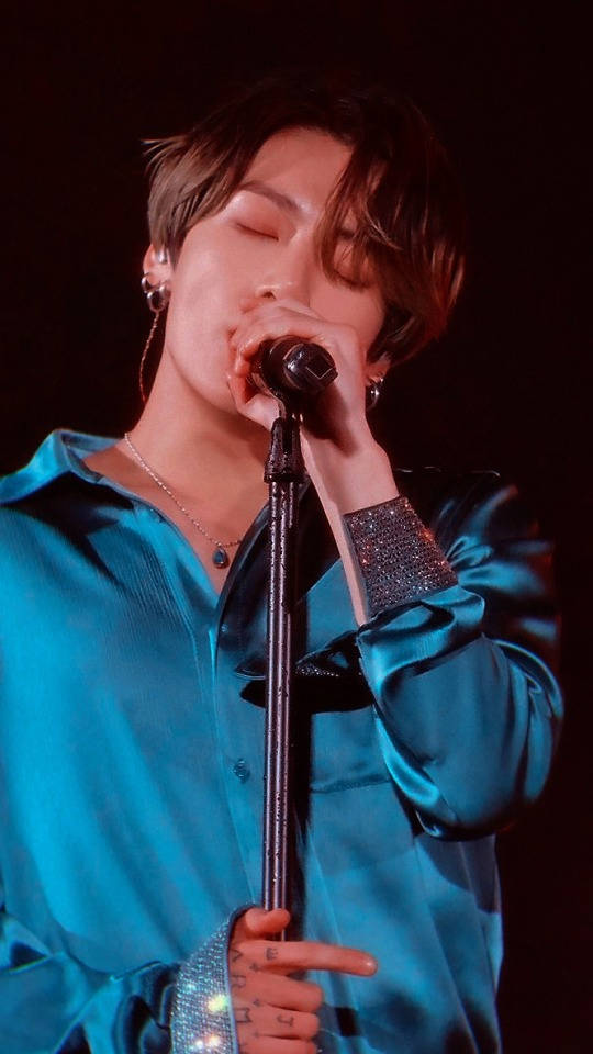 Jungkook 桌布 壁紙 — 一位表演者閉著眼睛對著麥克風唱歌，身穿閃亮的青綠色緞面襯衫，在舞台燈光下。
