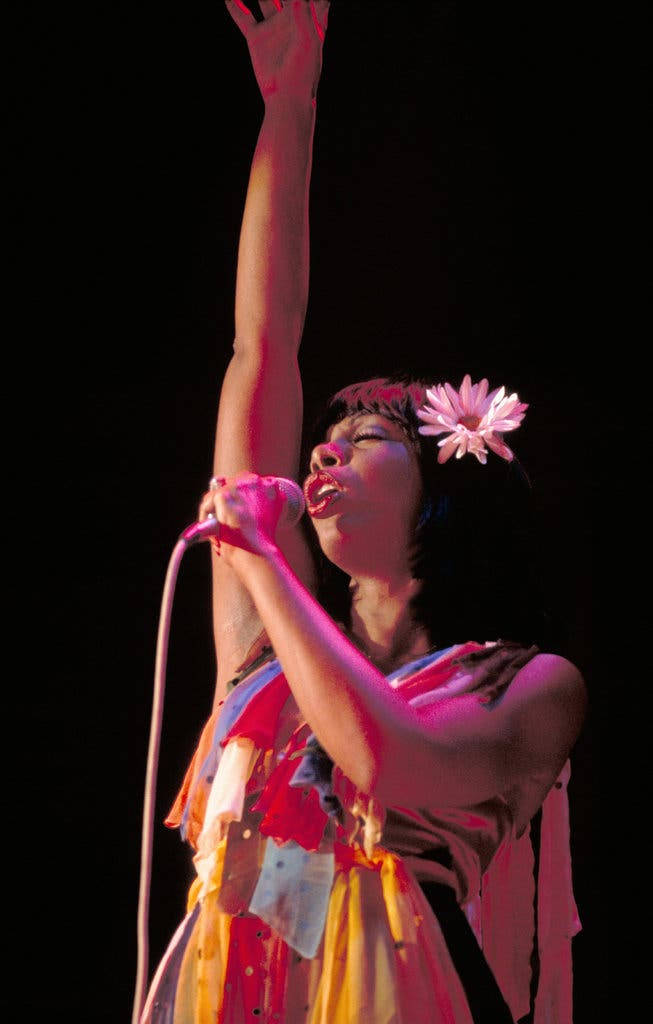Singing 桌布 壁紙 — 一位表演者頭髮上插著粉紅色花朵，在黑色背景的舞台燈光下熱情地對著麥克風唱歌。