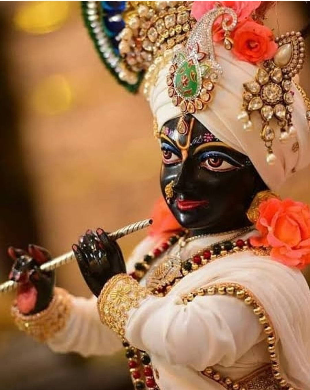 hindu iPhone 桌布 壁紙 — 一位裝飾華麗、深色皮膚的神祇，佩戴精緻珠寶和白色頭巾，在鮮豔的橙色玫瑰環繞下吹奏長笛。