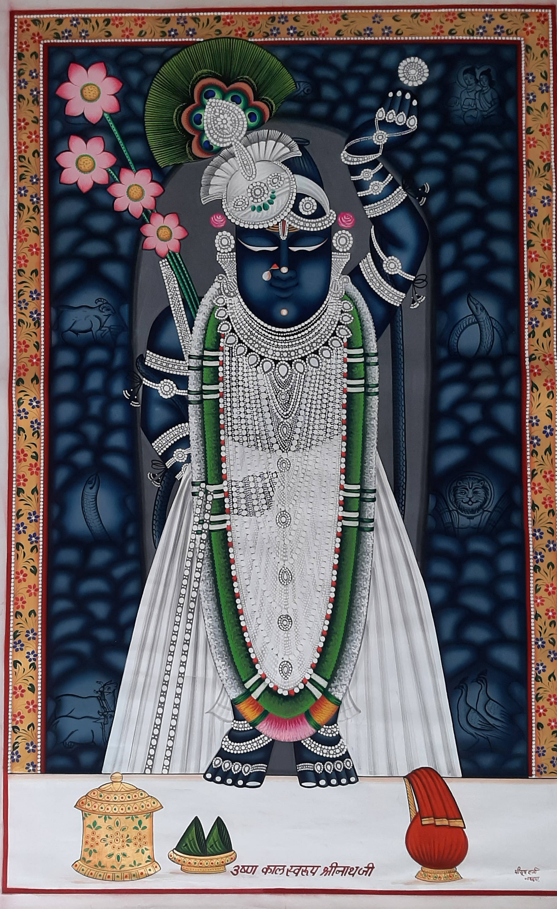 Shrinathji 桌布 壁紙 — 一位裝飾華麗的藍色神祇，佩戴精緻的銀飾和孔雀羽毛冠冕，站在深色圖案背景前，下方有花卉邊框和儀式物品。