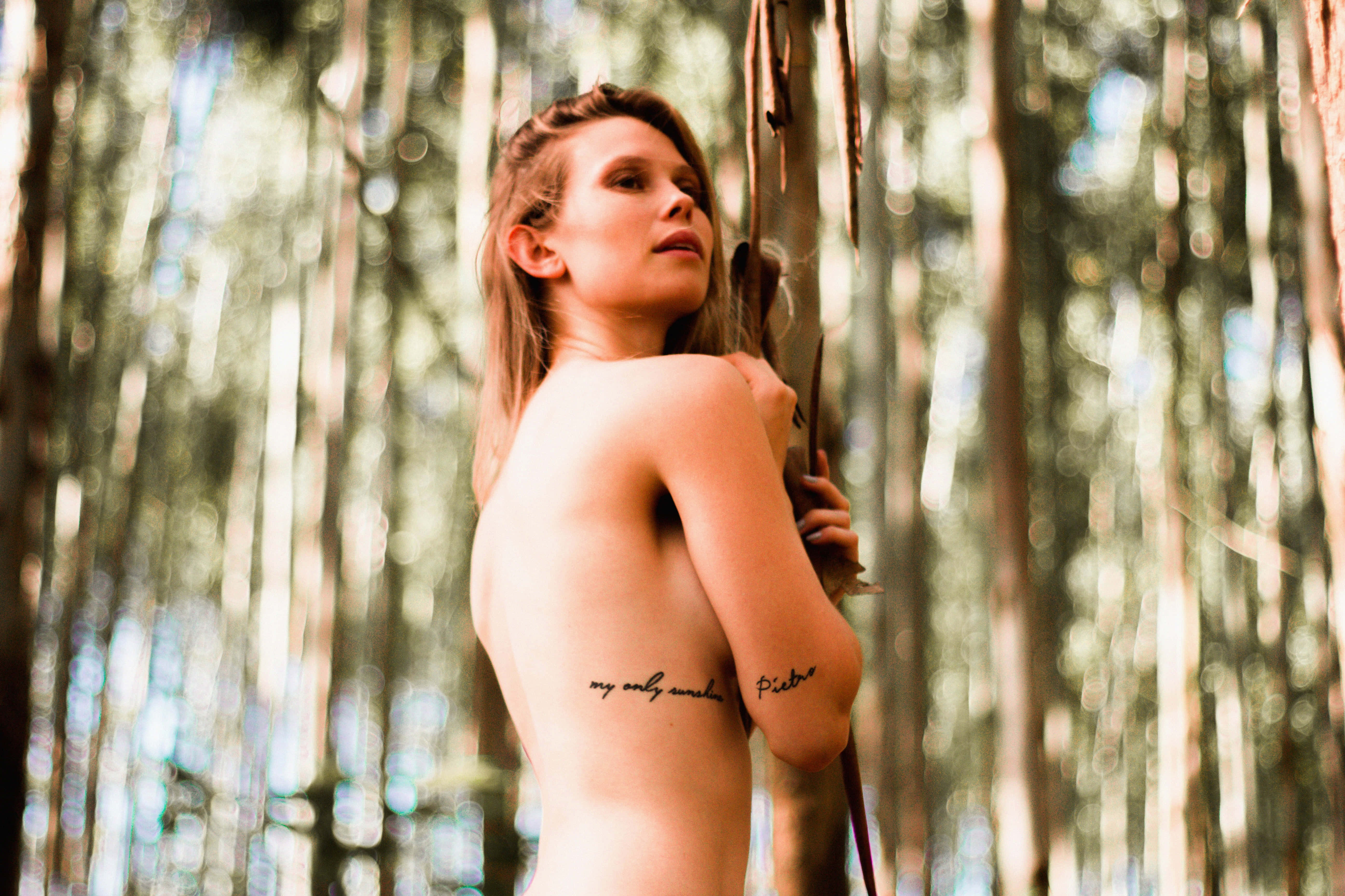 Tattoo 桌布 壁紙 — 一位裸體刺青女子站在陽光普照的森林中，手持樹枝，周圍是高聳的樹木，光線斑駁。