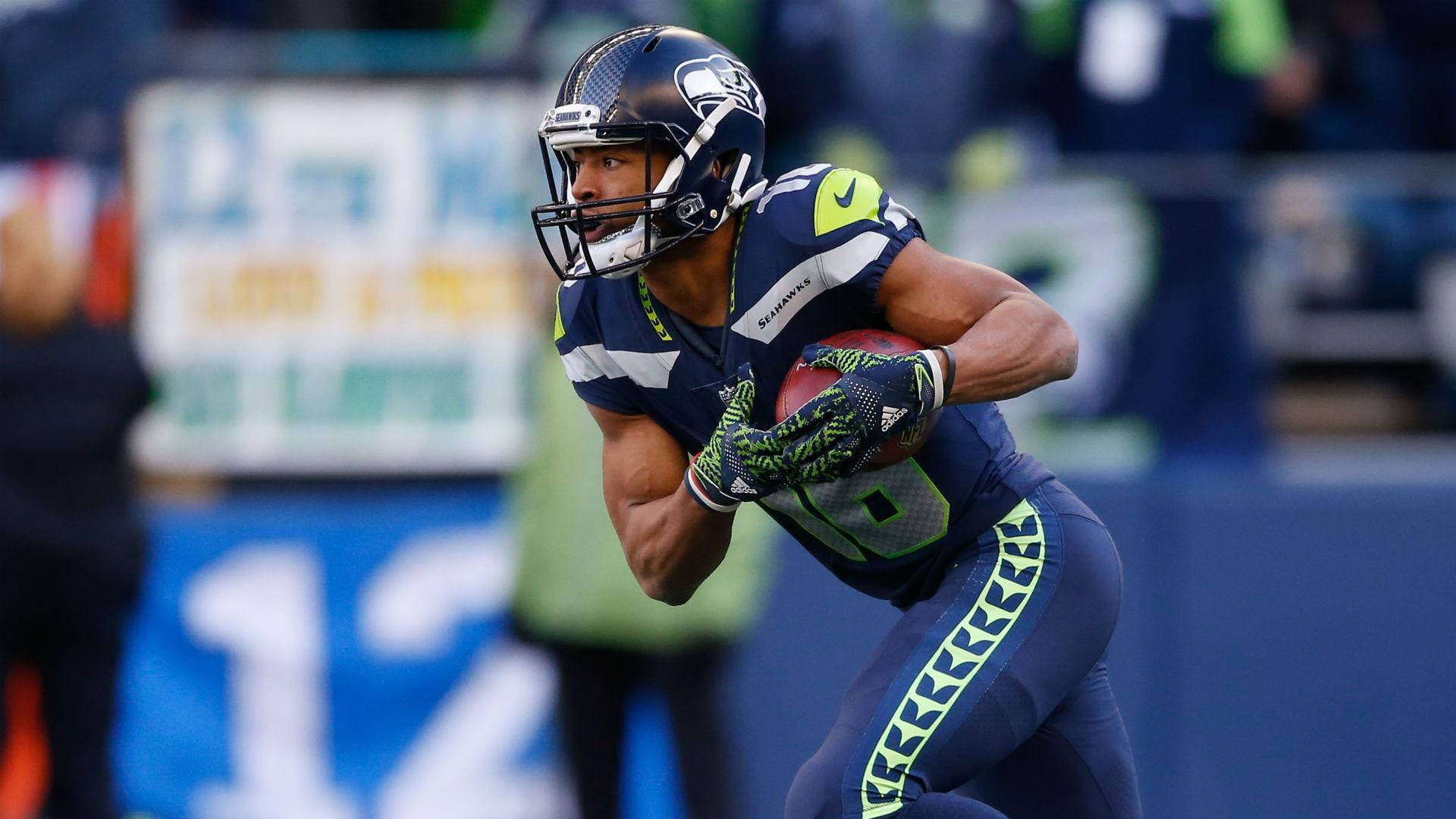 Seattle Seahawks 桌布 壁紙 — 一位西雅圖海鷹隊的美式足球員身穿海軍藍和螢光綠色制服，在日間比賽中持球衝刺。