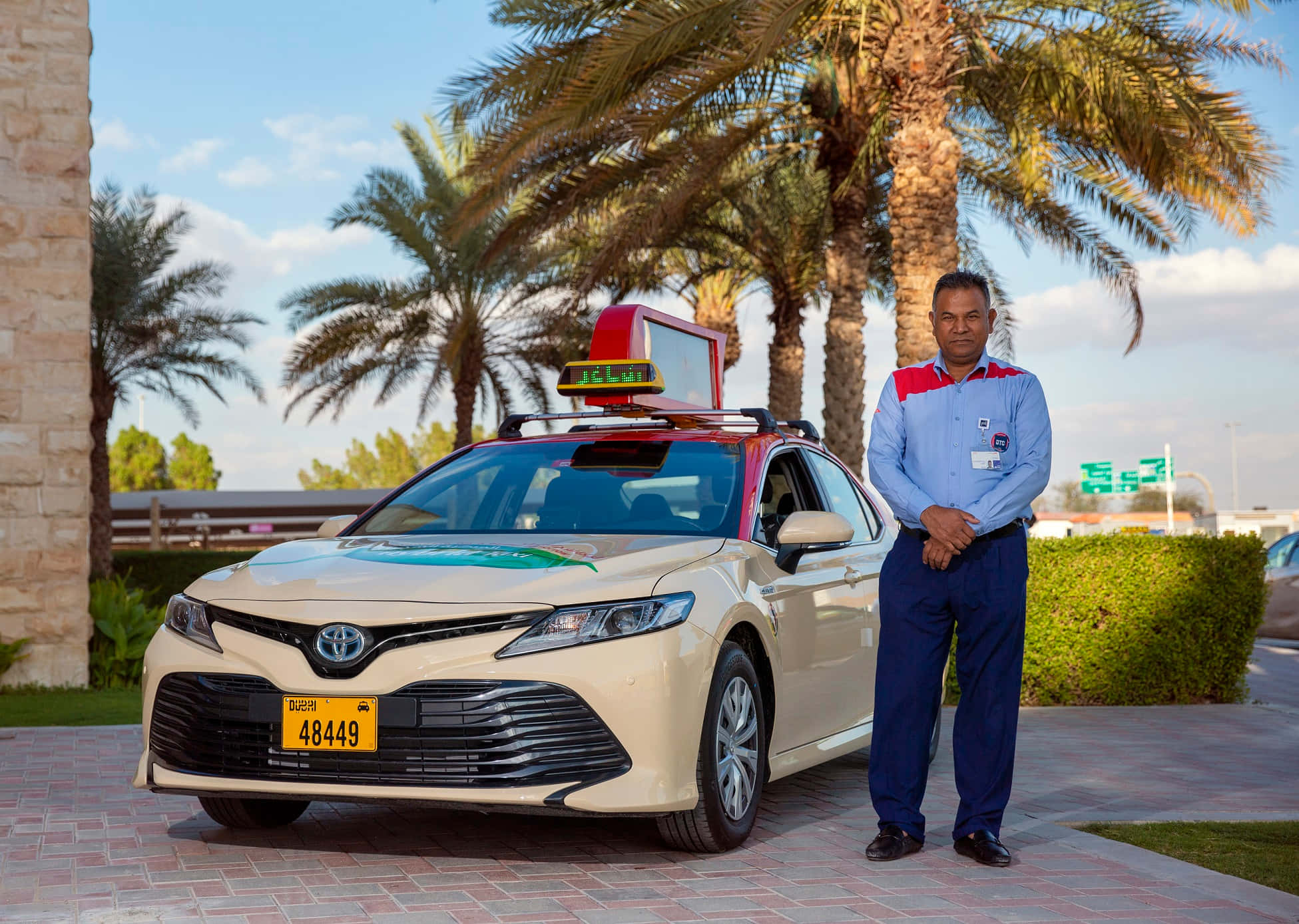 taxi 桌布 壁紙 — 一位計程車司機在陽光明媚的沙漠環境中，自豪地站在棕櫚樹下他的奶油色豐田Camry計程車旁邊。