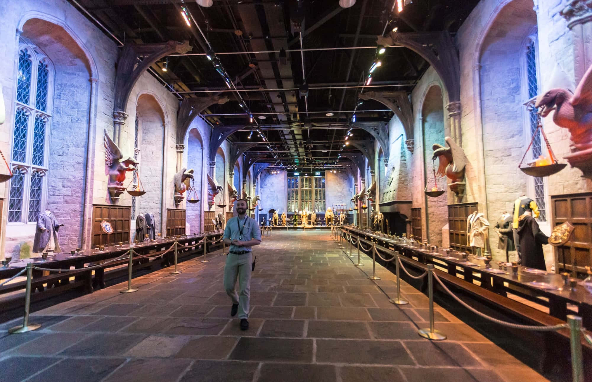 The Hogwarts Great Hall 桌布 壁紙 — 一位訪客走過宏偉、燈光昏暗的霍格華茲大禮堂，石拱門和魔法展示沿著大廳排列。