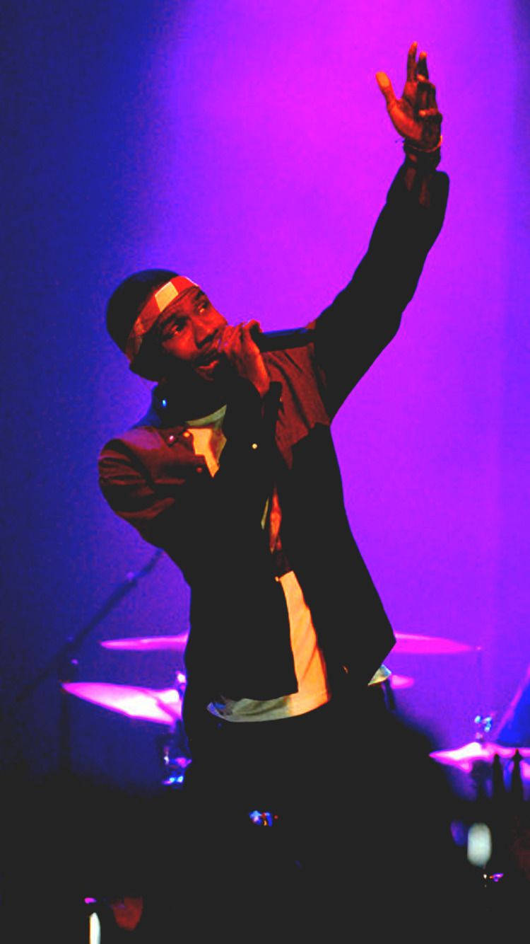 Frank Ocean 桌布 壁紙 — 一位說唱歌手在充滿活力的紫色和藍色燈光下，以一隻手臂舉起的動態姿勢在舞台上表演。