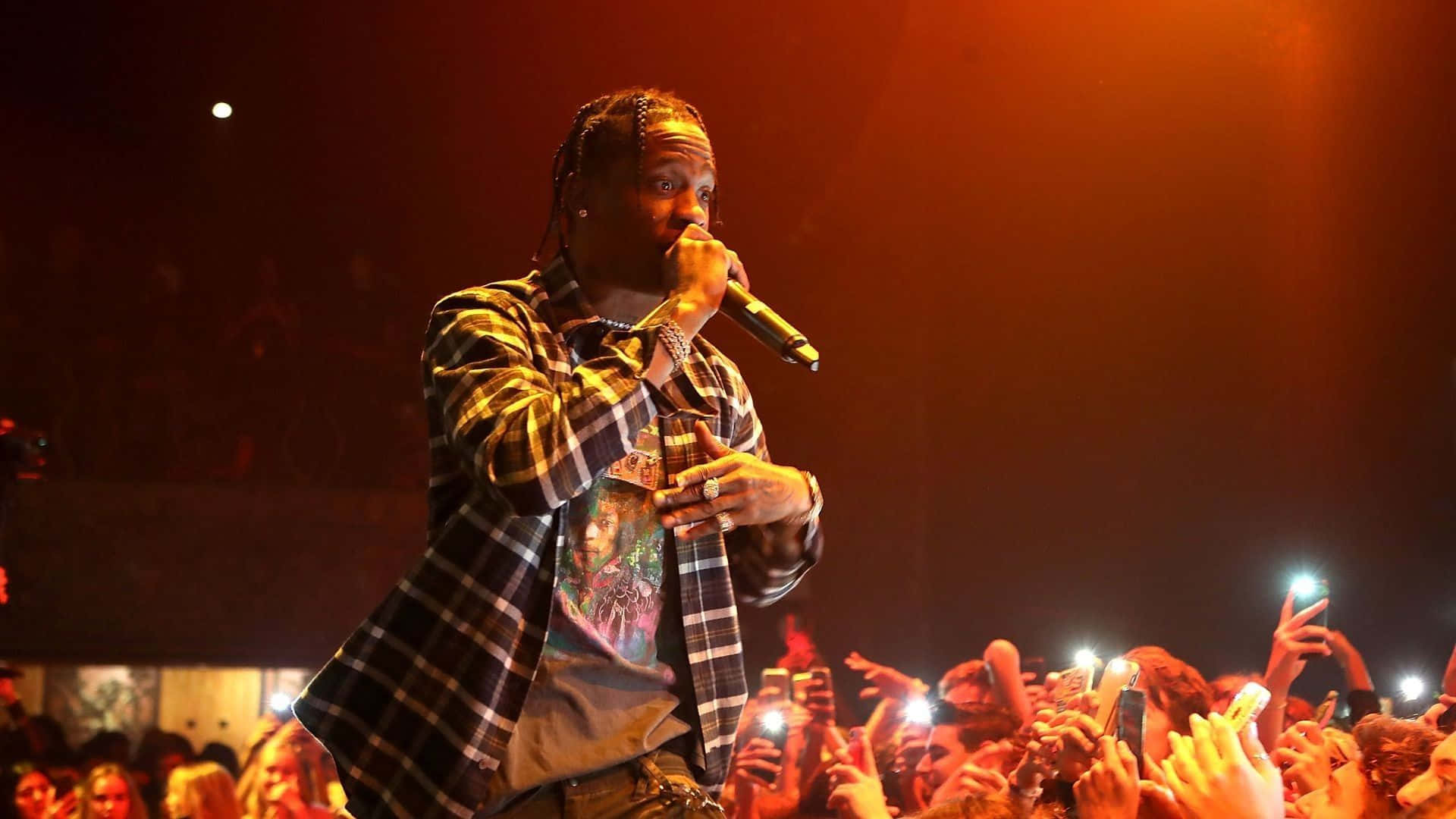 Travis Scott Concert 桌布 壁紙 — 一位說唱歌手在溫暖的橙色燈光下於舞台上表演，充滿活力的觀眾舉起發光的手機螢幕。