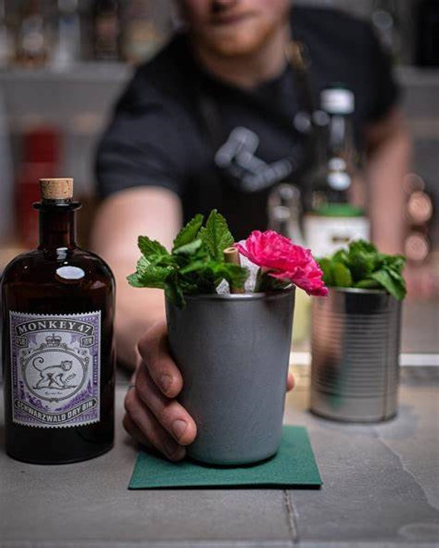 cocktail 桌布 壁紙 — 一位調酒師拿著一個灰色錫杯，裡面裝滿了琴酒雞尾酒，飾以薄荷和粉紅花朵，旁邊放著一瓶Monkey 47琴酒。