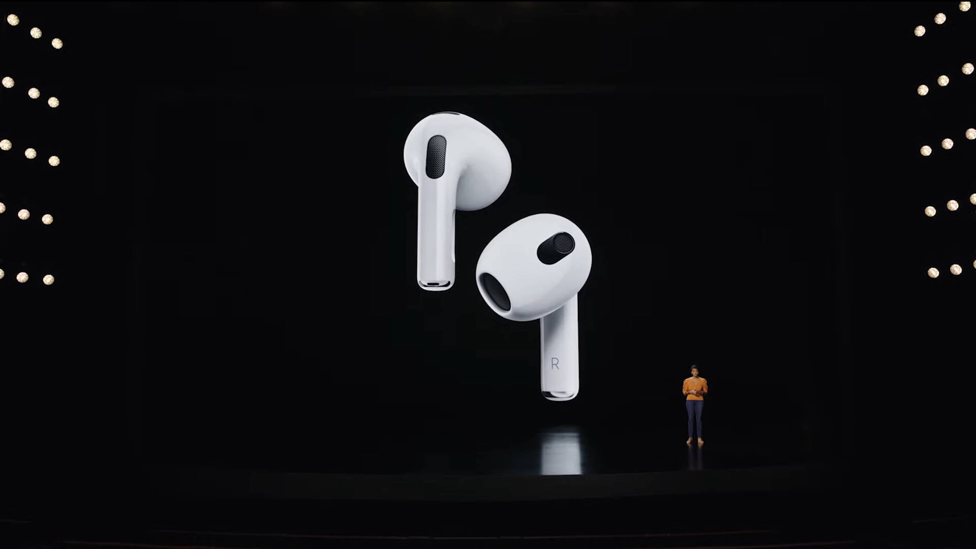 AirPods 桌布 壁紙 — 一位講者站在黑暗的舞台上，面前是一個巨大的螢幕，顯示兩側帶有發光燈光的浮動白色無線耳塞。