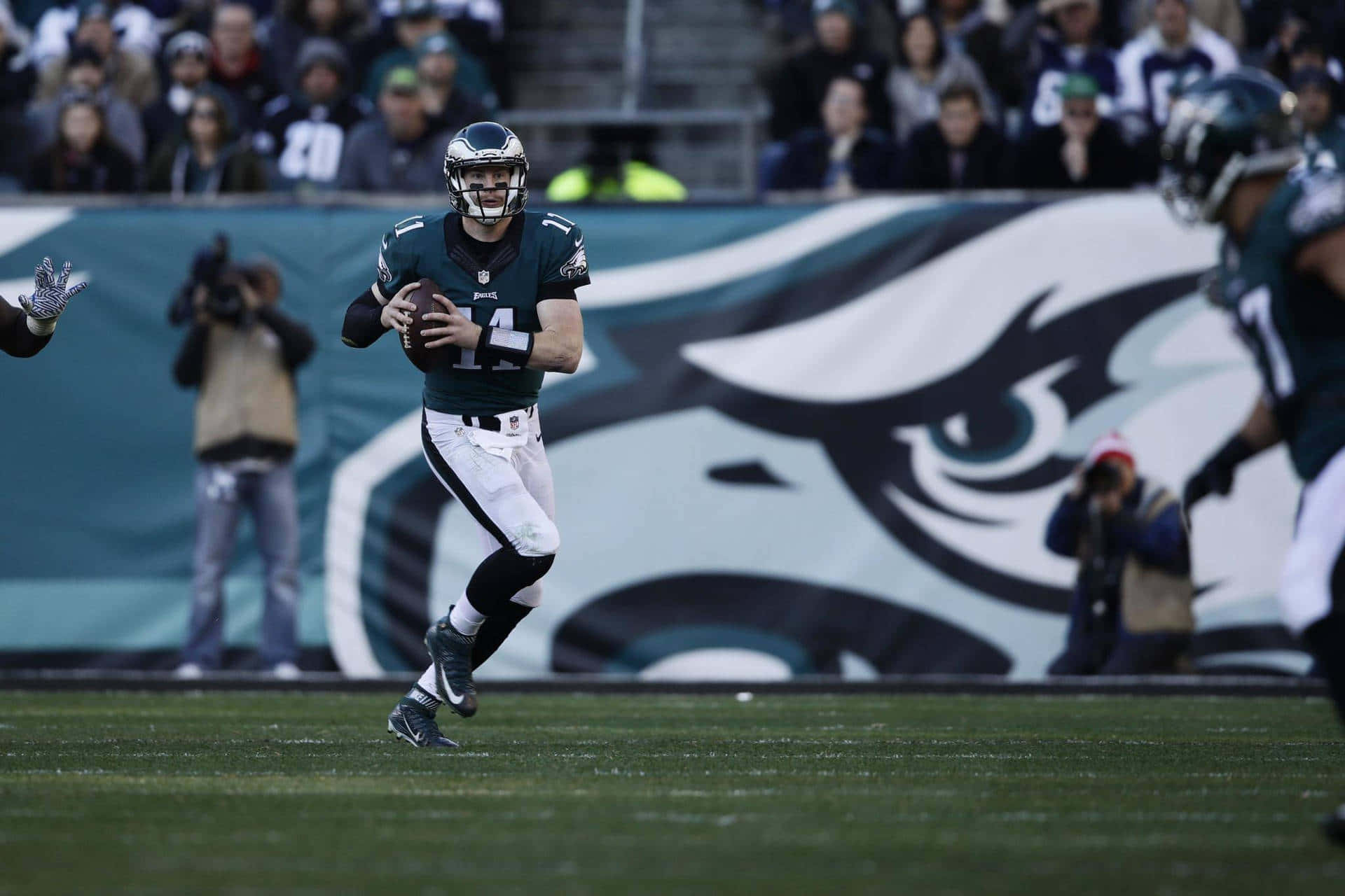 Eagles Football 桌布 壁紙 — 一位費城老鷹隊的四分衛身穿綠白相間的球衣，在草地場上比賽時帶著橄欖球奔跑，背景有球迷和球隊標誌。