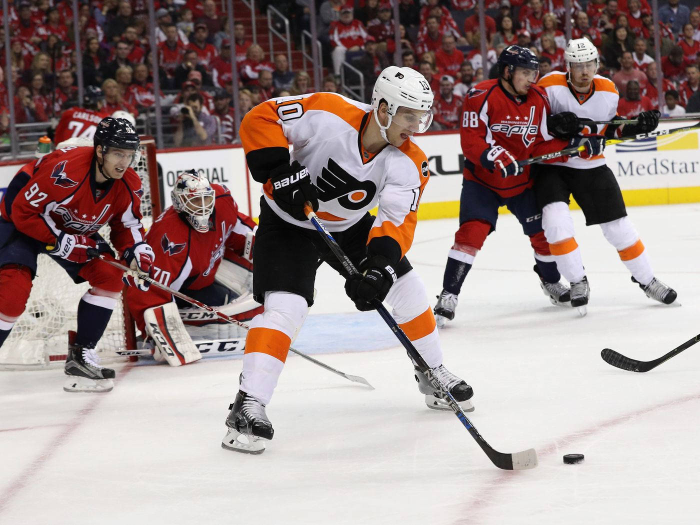 Philadelphia Flyers 桌布 壁紙 — 一位費城飛人隊球員身穿白色和橙色球衣，在華盛頓首都隊後衛的壓迫下控制冰球，進行一場激烈的NHL比賽。