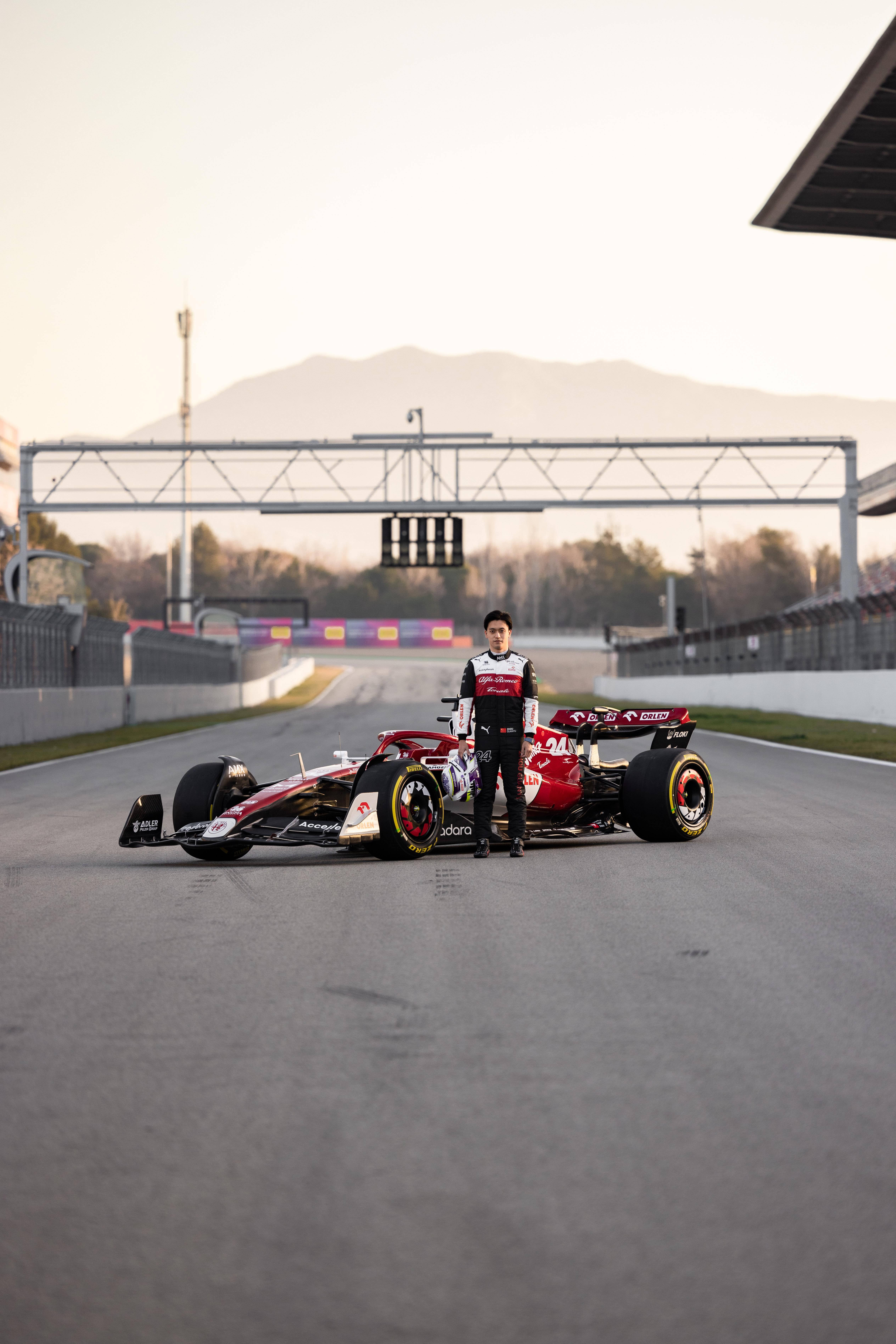 Racing 桌布 壁紙 — 一位賽車手自信地站在一輛紅黑相間的一級方程式賽車旁，背景是空蕩蕩的賽道和金色時刻的山巒。
