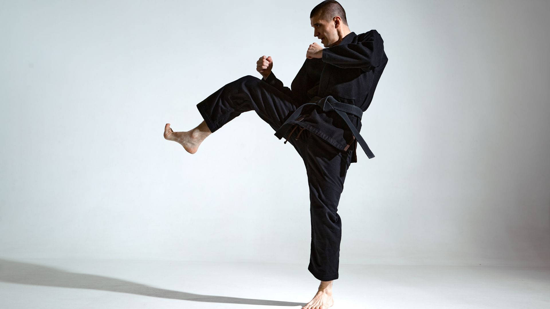 martial arts 桌布 壁紙 — 一位赤腳的武術家穿著黑色道服，在極簡的白色工作室背景下執行高踢腿。