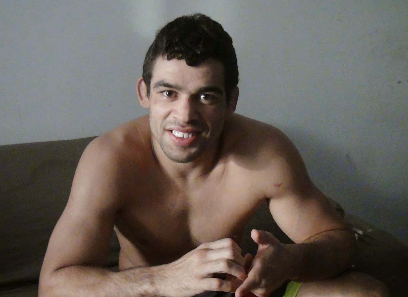 UFC 桌布 壁紙 — 一位赤膊、肌肉發達、深色頭髮的男子坐在光線昏暗的房間的沙發上，直視鏡頭微笑。