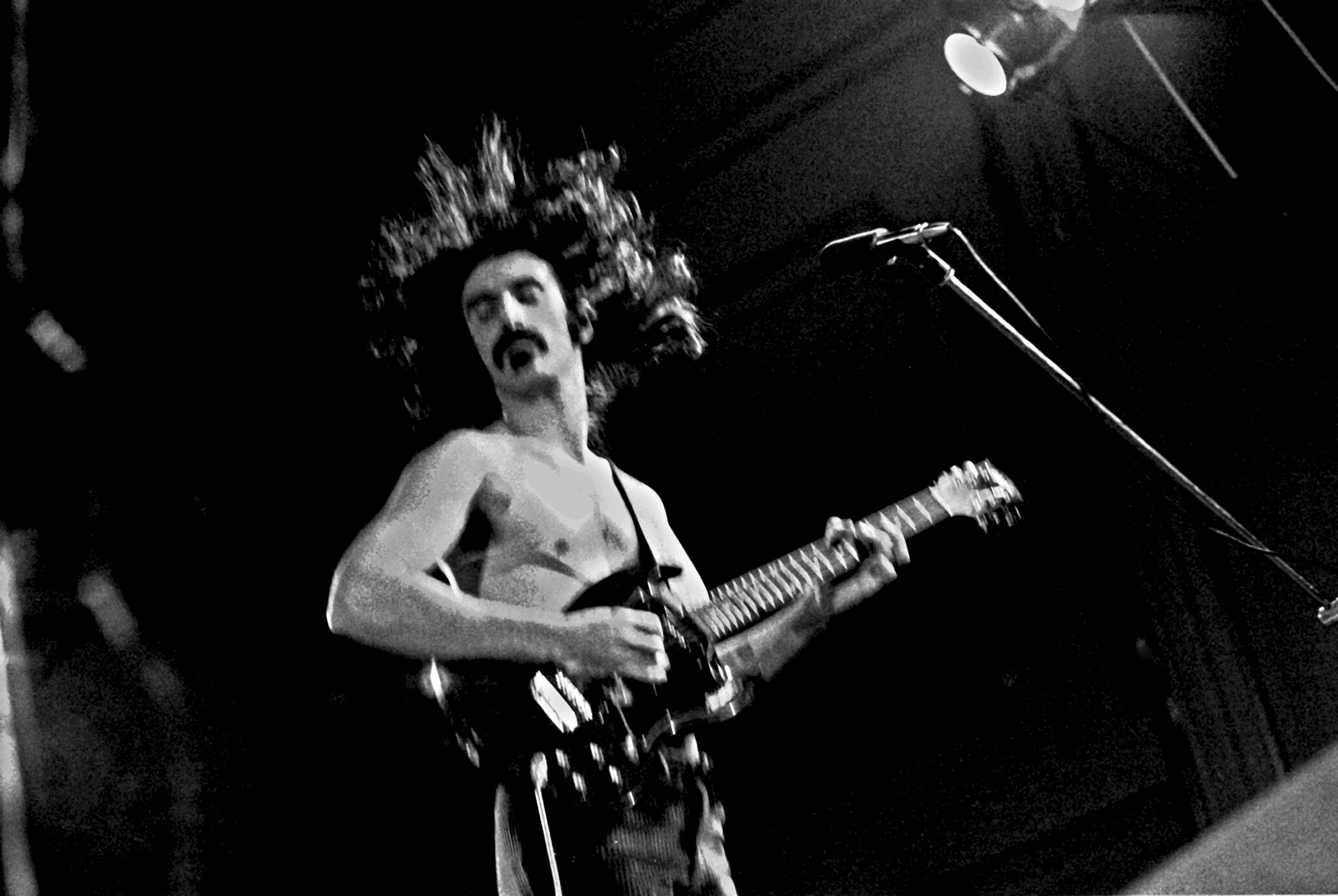 Zappa 桌布 壁紙 — 一位赤膊上陣、頭髮狂野的音樂家在黑暗的舞台上，在單一聚光燈下彈奏電吉他。