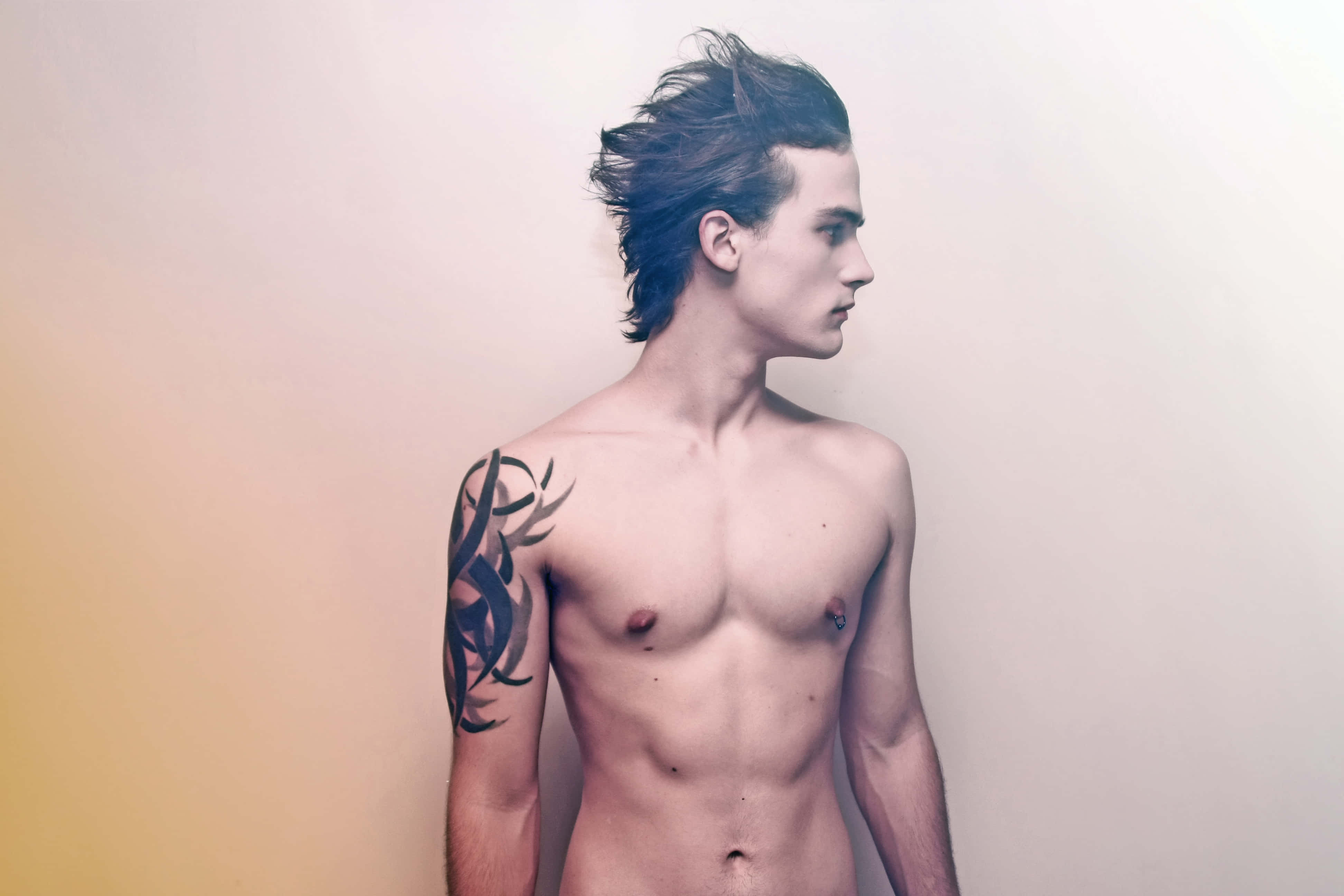 Hot Guy 桌布 壁紙 — 一位赤膊的深色頭髮年輕男子，左臂上有部落圖騰刺青，側身站在柔和的漸變背景前。