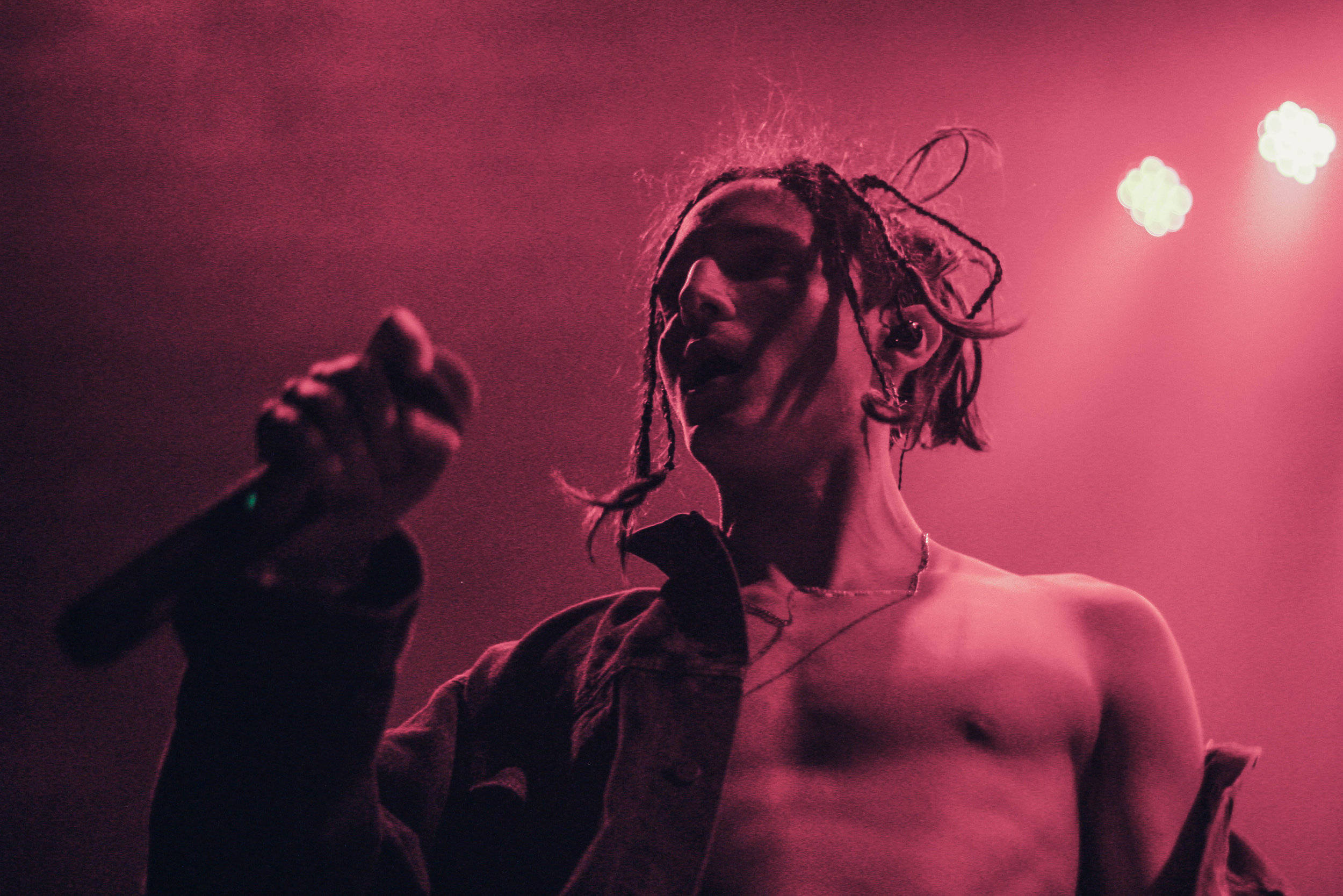 Chase Atlantic 桌布 壁紙 — 一位赤裸上身、留著辮子頭的表演者在強烈的粉紅色舞台燈光下，熱情地對著麥克風唱歌。