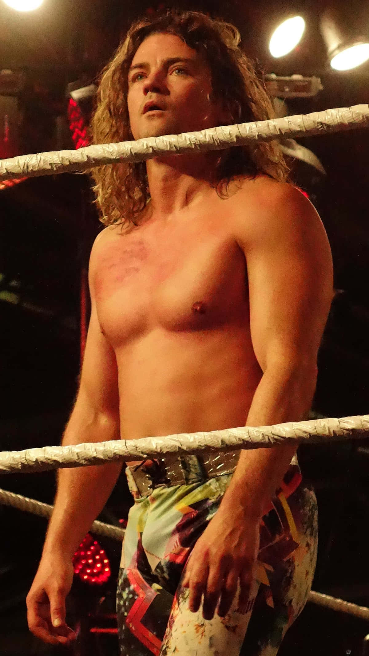 WWE 桌布 壁紙 — 一位赤裸上身、長著捲曲長髮的摔角手，在明亮的聚光燈下站在擂台上，神情專注。