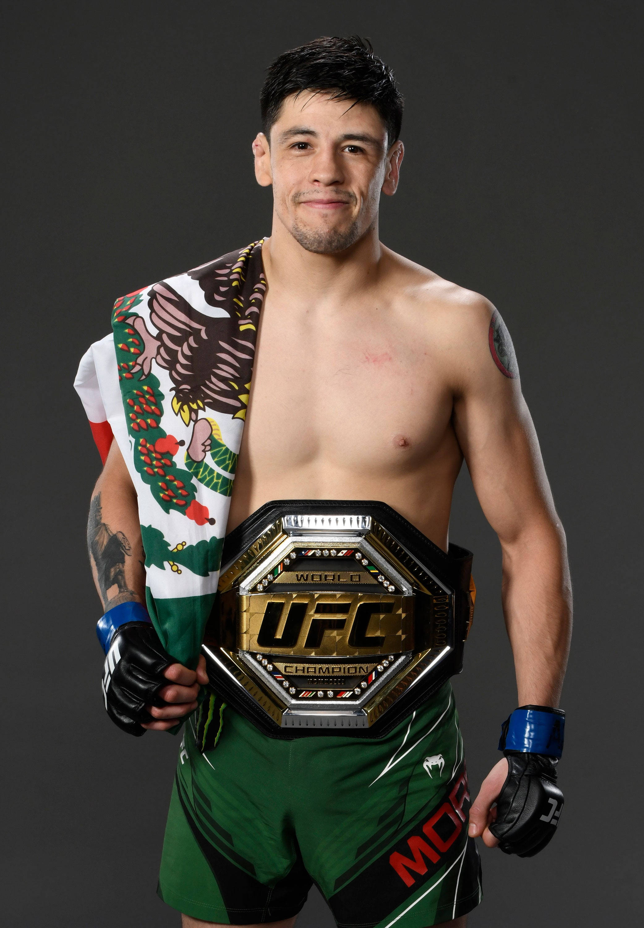 Brandon Moreno 桌布 壁紙 — 一位赤裸上身的UFC冠軍自豪地舉起他的冠軍腰帶，肩上披著墨西哥國旗。