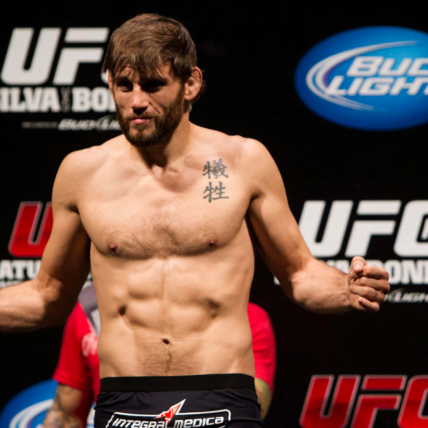 UFC 桌布 壁紙 — 一位赤裸上身的UFC拳手，胸前有刺青，在明亮的燈光下自信地站在稱重活動中。