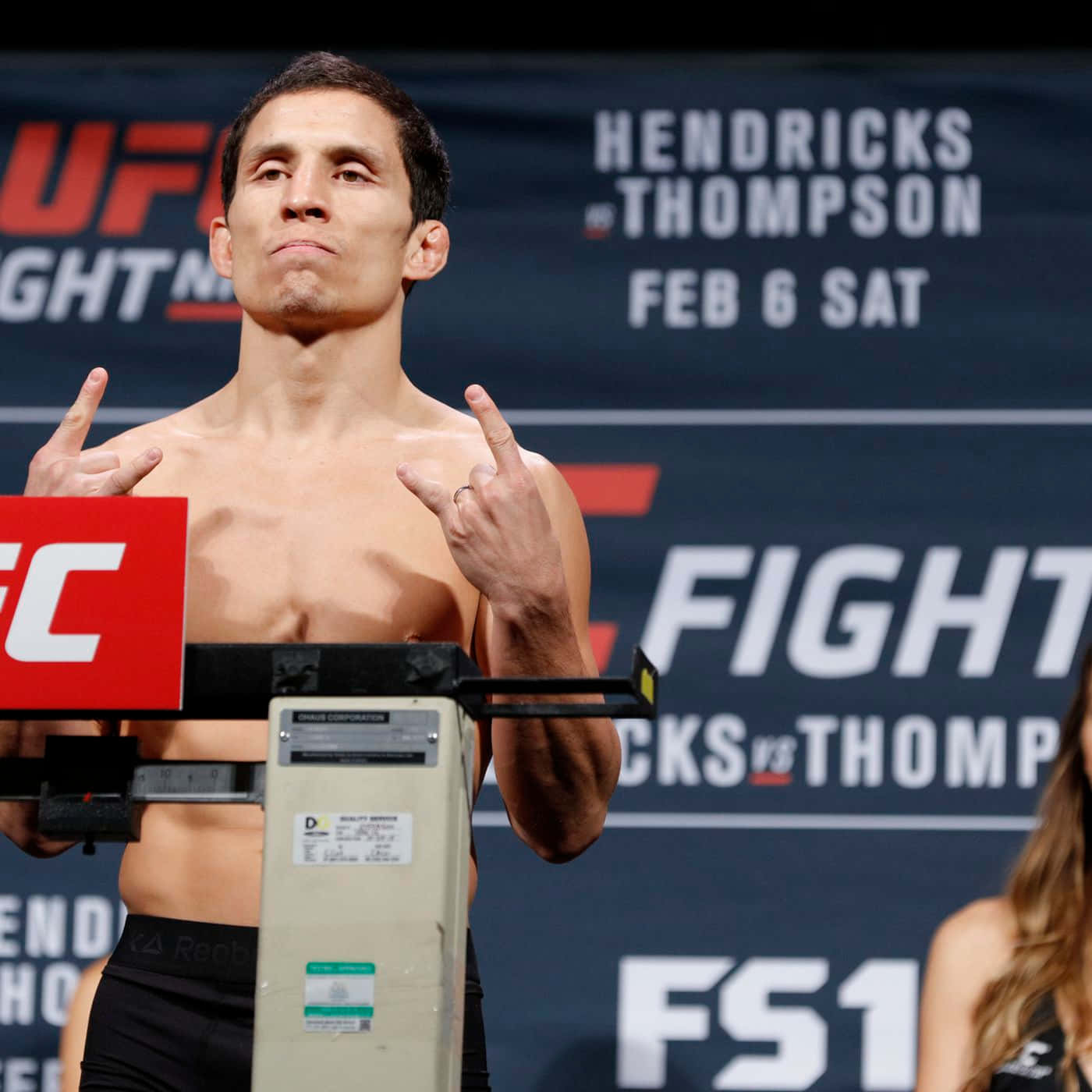 UFC 桌布 壁紙 — 一位赤裸上身的UFC拳手在稱重活動中站在體重磅上，做出搖滾手勢，表情專注。