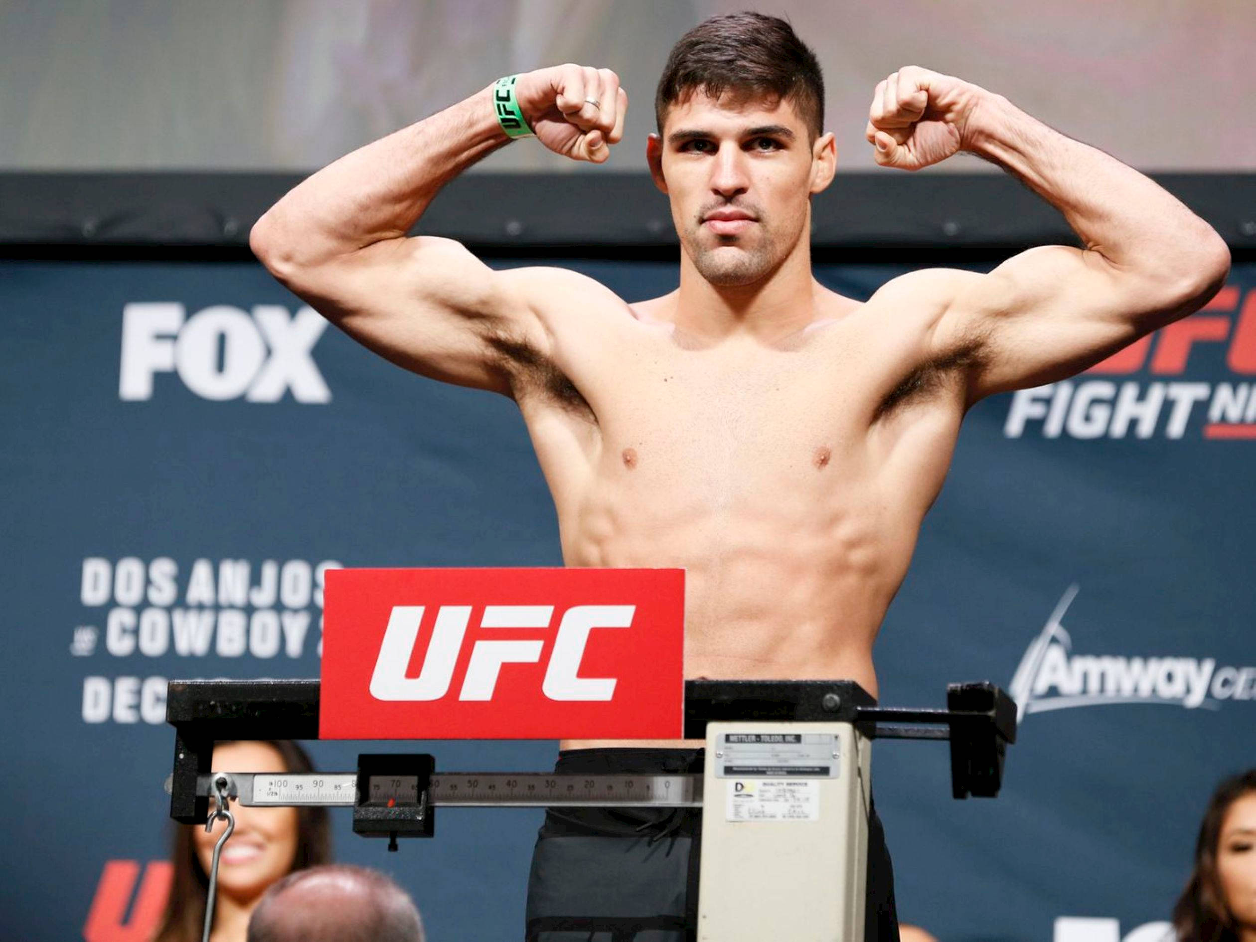 MMA 桌布 壁紙 — 一位赤裸上身的UFC拳手在稱重活動中，在體重磅上展示他肌肉發達的手臂，背後有FOX和Amway的品牌標誌。
