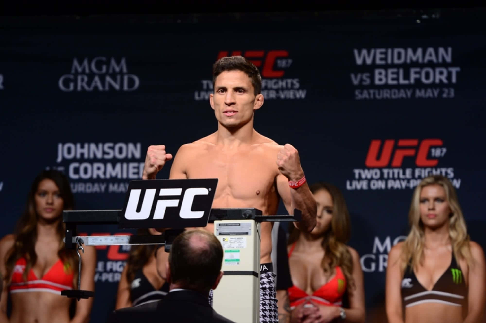 MMA 桌布 壁紙 — 一位赤裸上身的UFC拳手自信地站在體重磅上，舉起拳頭，身後有身穿比基尼的擂台女郎在稱重活動中。