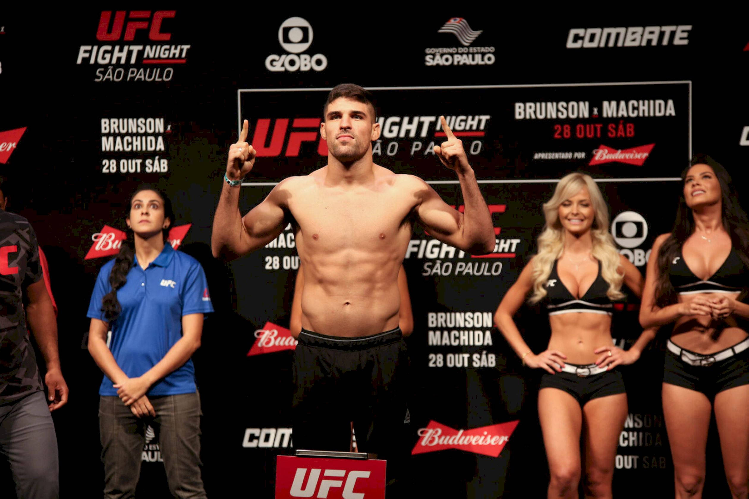 MMA 桌布 壁紙 — 一位赤裸上身的UFC拳手自信地站在體重磅上，進行稱重活動，附近有兩位身穿比基尼的擂台女郎和官員。