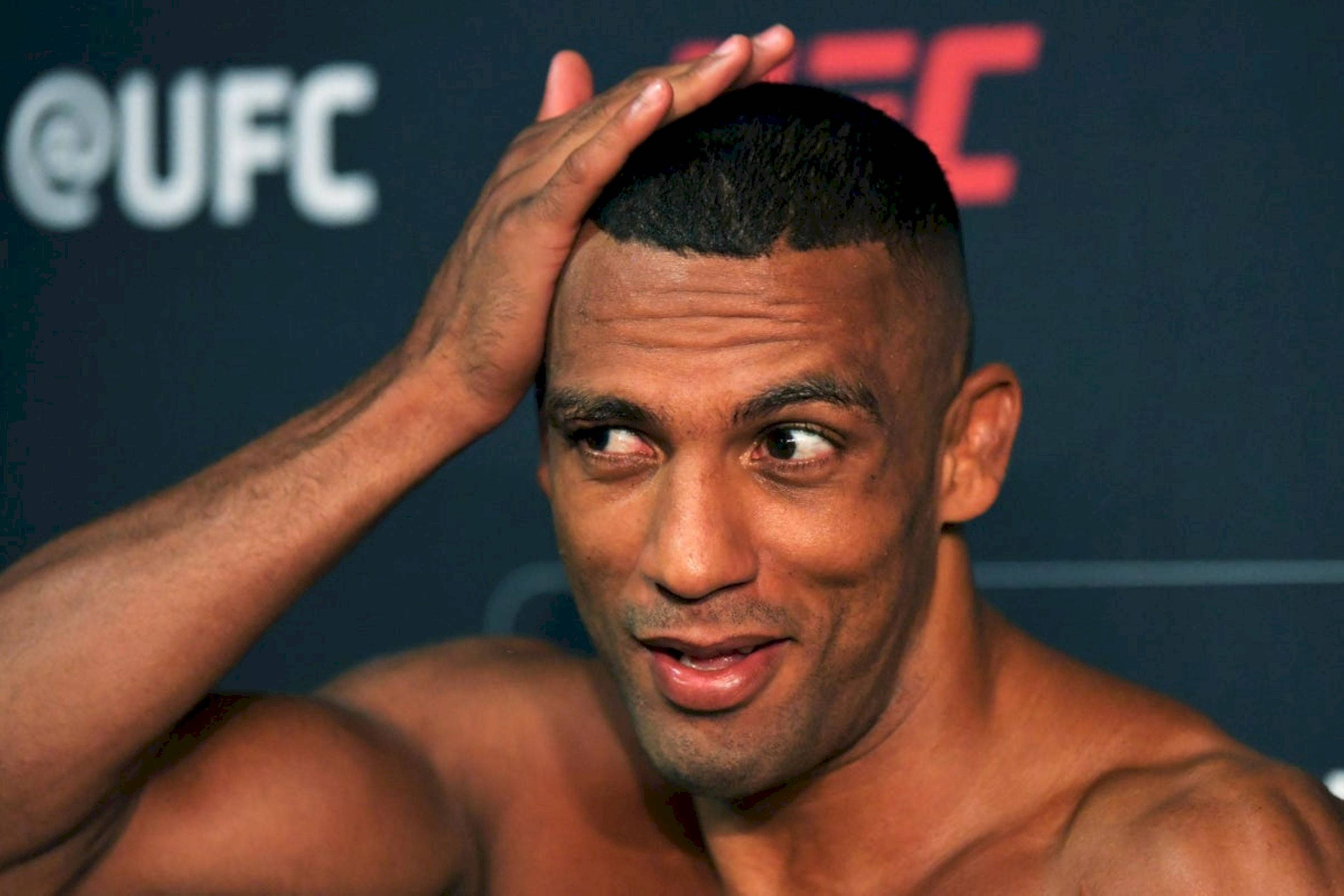 MMA 桌布 壁紙 — 一位赤裸上身的UFC拳擊手將手放在頭上，在品牌背景前側視，露出頑皮的微笑。