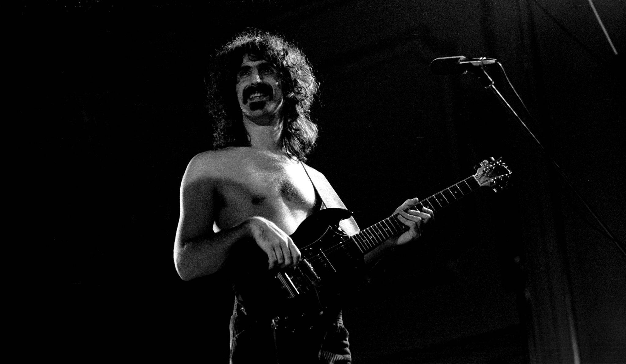 Zappa 桌布 壁紙 — 一位赤裸上身的吉他手，留著長捲髮和鬍子，在舞台燈光下熱情地彈奏電吉他。