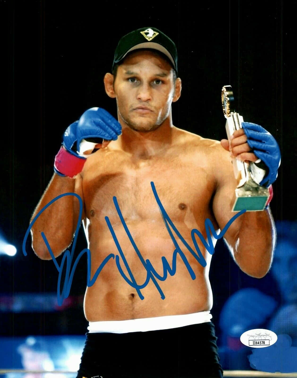 Boxing 桌布 壁紙 — 一位赤裸上身的拳擊手，戴著藍色手套和帽子，在黑暗的競技場背景下，神情嚴肅地舉著獎盃。