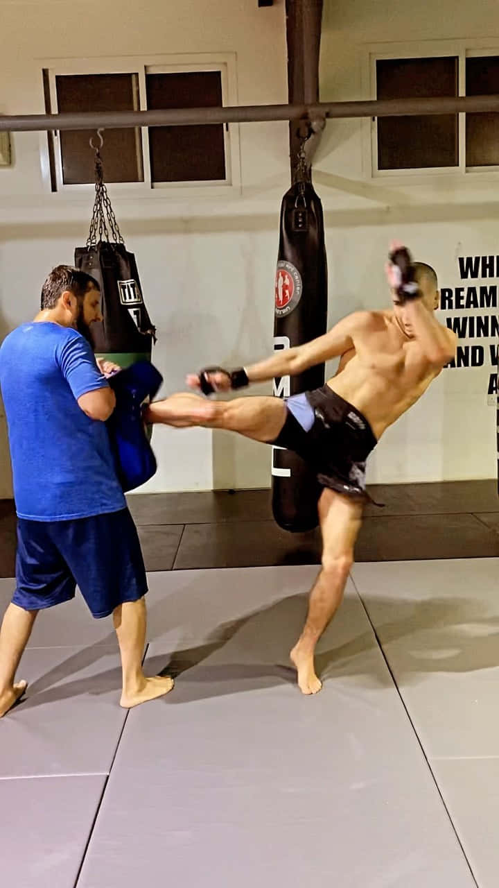 MMA 桌布 壁紙 — 一位赤裸上身的武術家在健身房中，對著教練穩穩抓住的重沙包施展高踢腿。