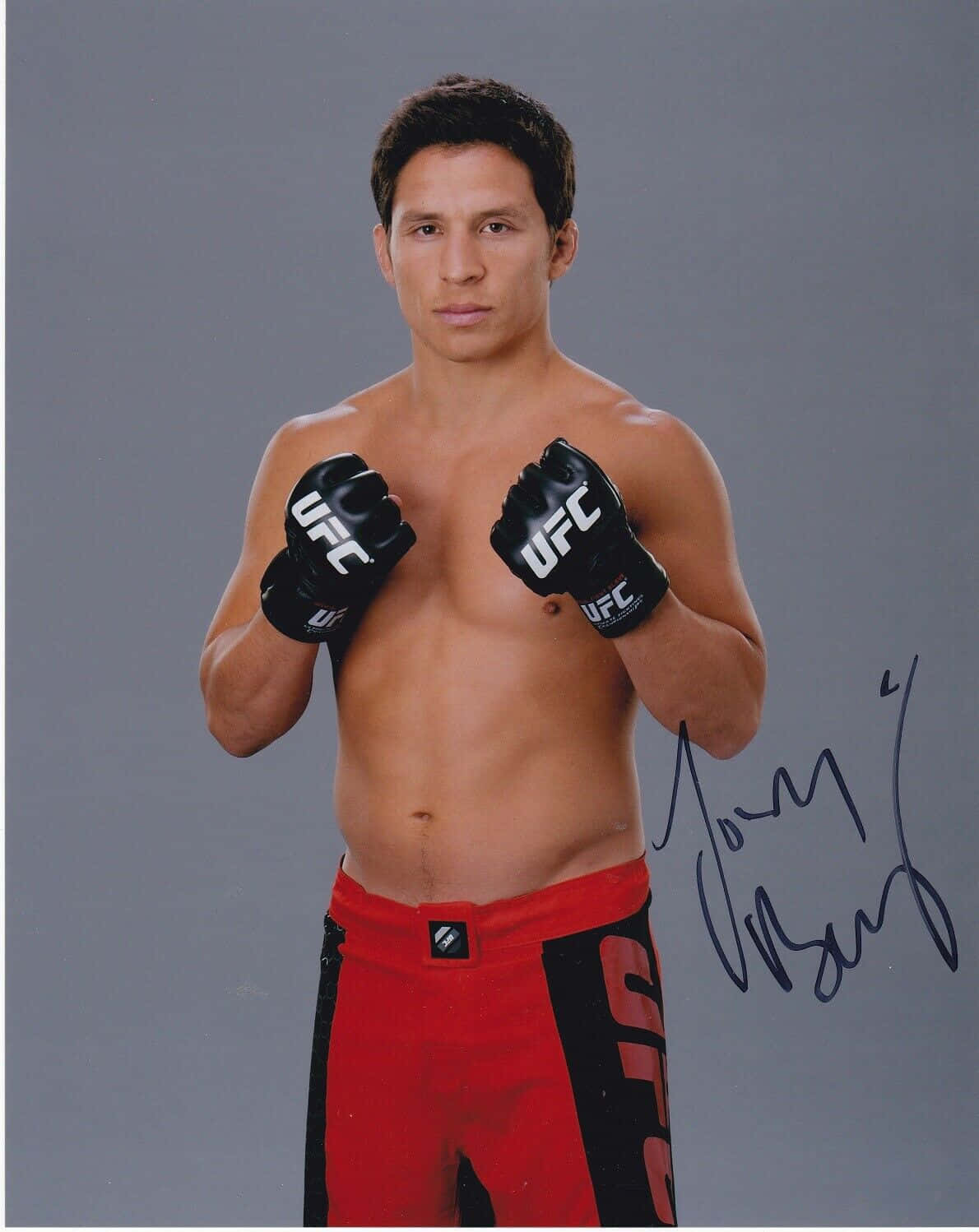 MMA 桌布 壁紙 — 一位赤裸上身的男性混合武術家，穿著紅色短褲和黑色UFC手套，在純灰色工作室背景前自信地擺姿勢，他的簽名清晰可見。