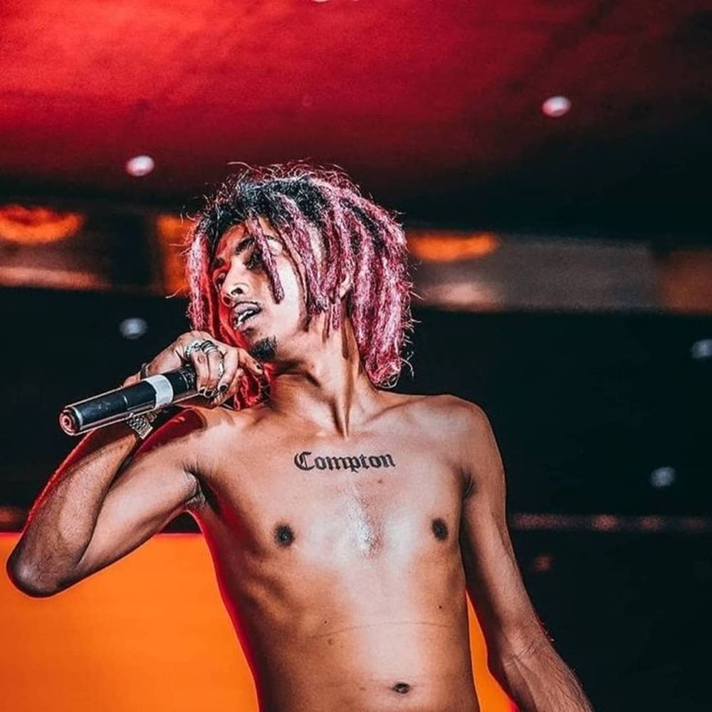說唱歌手 桌布 壁紙 — 一位赤裸上身的男性表演者，留著粉紅色辮子，在紅色舞台燈光下對著麥克風唱歌，胸前紋有「Compton」字樣。