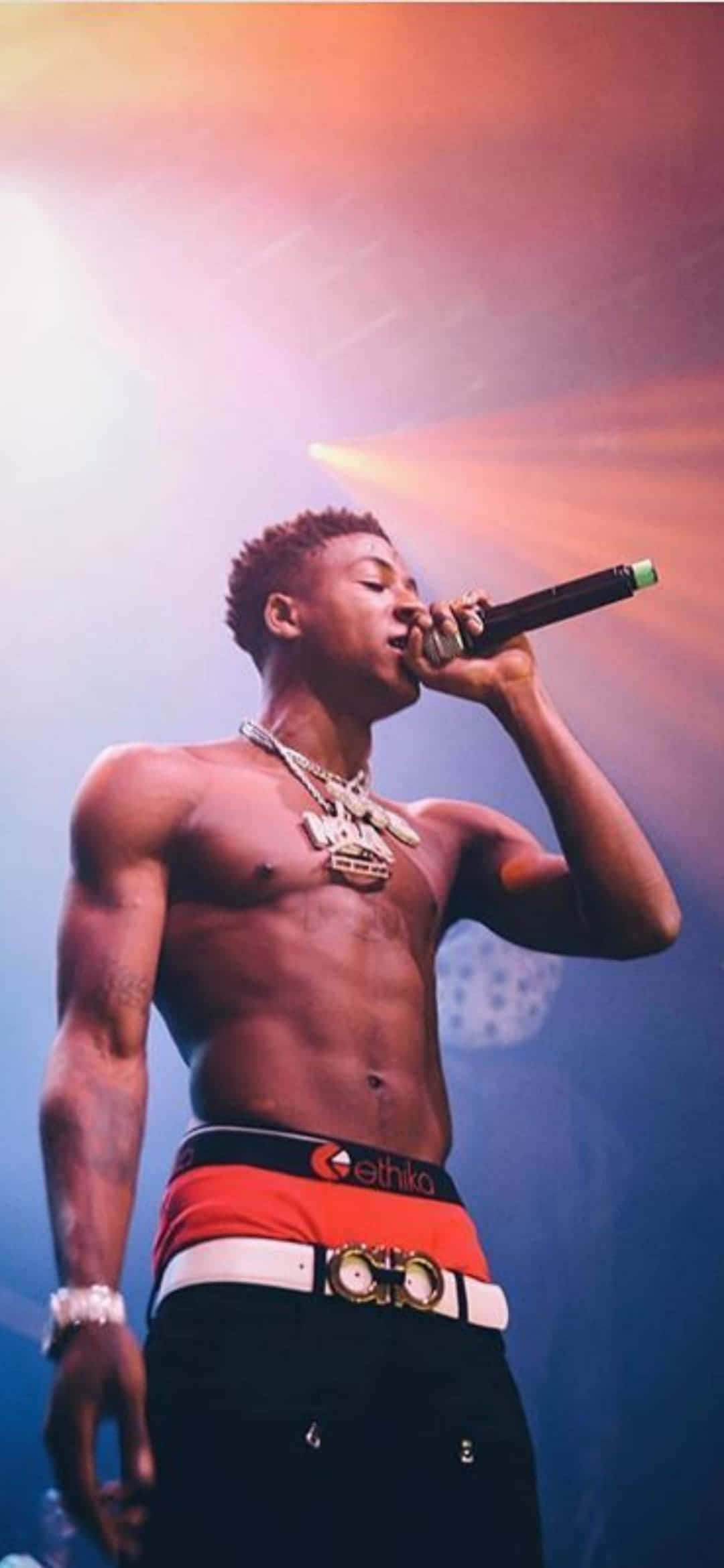 Youngboy 桌布 壁紙 — 一位赤裸上身的男饒舌歌手戴著金鏈，在充滿活力的舞台燈光下對著麥克風熱情表演。