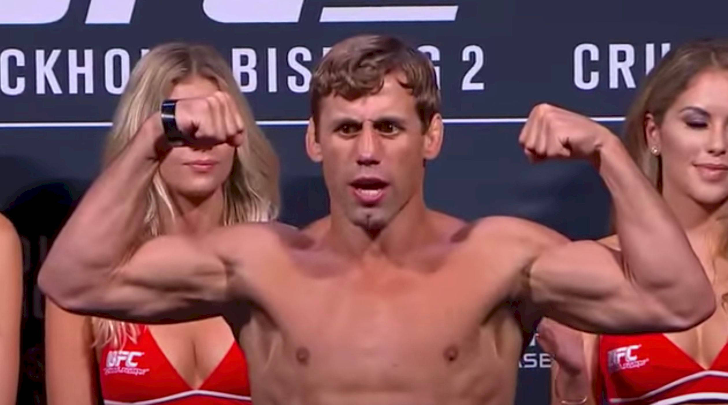 MMA 桌布 壁紙 — 一位赤裸上身的肌肉發達拳手在UFC稱重活動中彎曲雙臂，身旁有兩位身穿紅色服裝的女性。