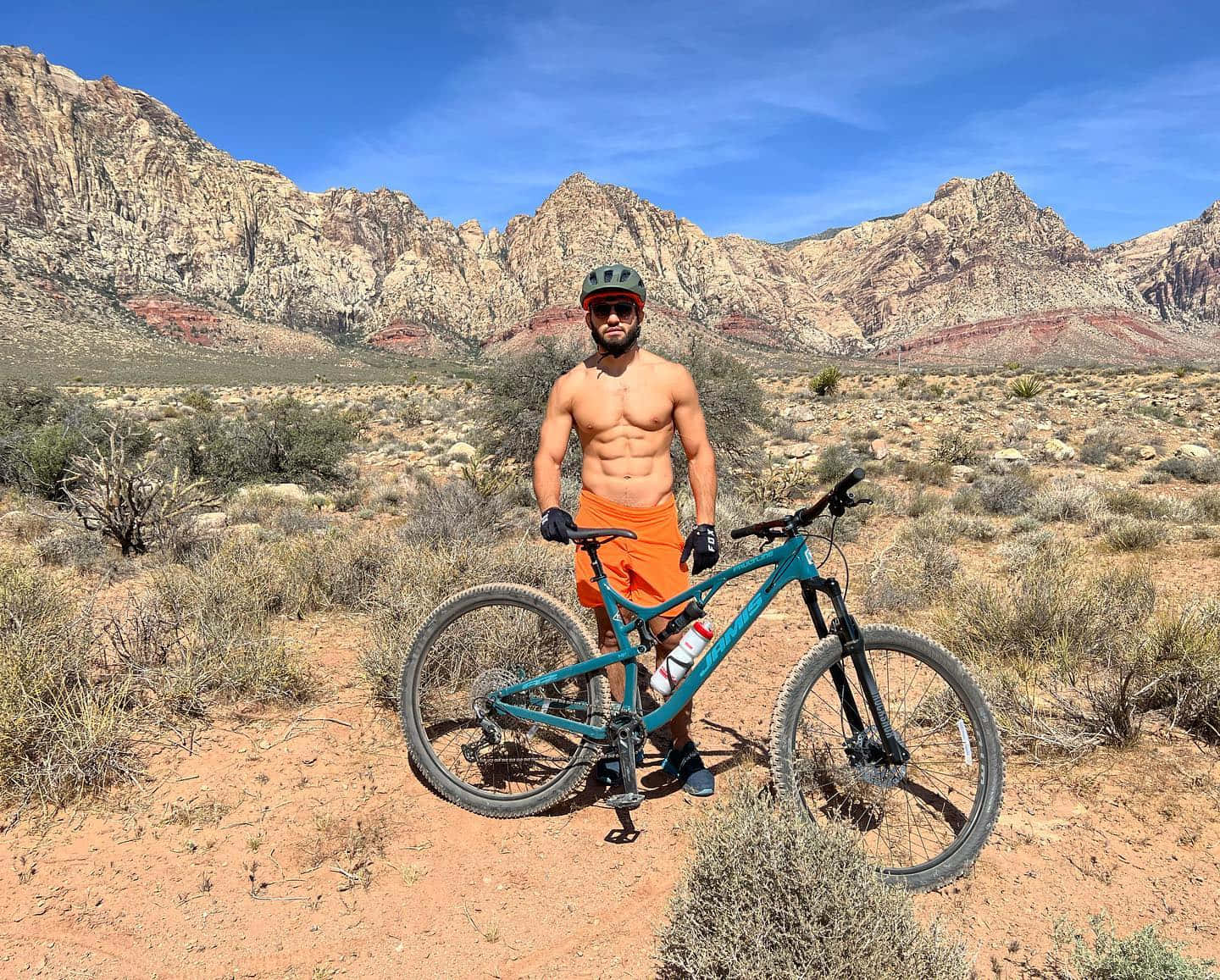 Athlete 桌布 壁紙 — 一位赤裸上身的自行車手，穿著橙色短褲和頭盔，自豪地站在他的青綠色登山車旁，背景是崎嶇的沙漠山脈，天空清澈湛藍。